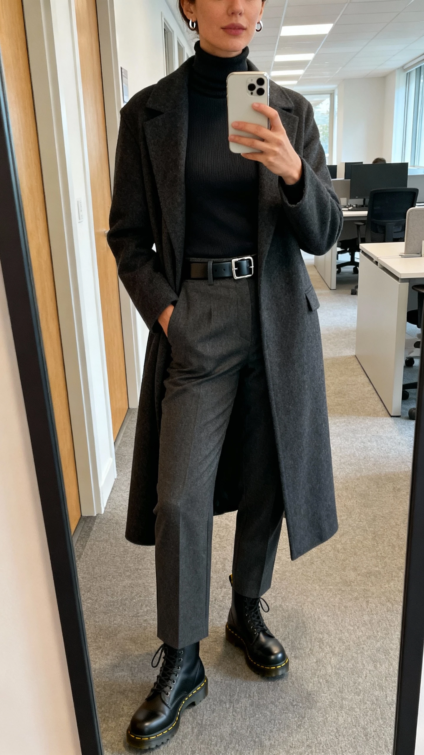 iPhone mirror selfie of a woman wearing tailored trousers, fitted turtleneck, longline coat, sleek belt, Doc Martens, simple earrings, face not visible, office hallway mirror, morning natural light, iPhone photo quality.
