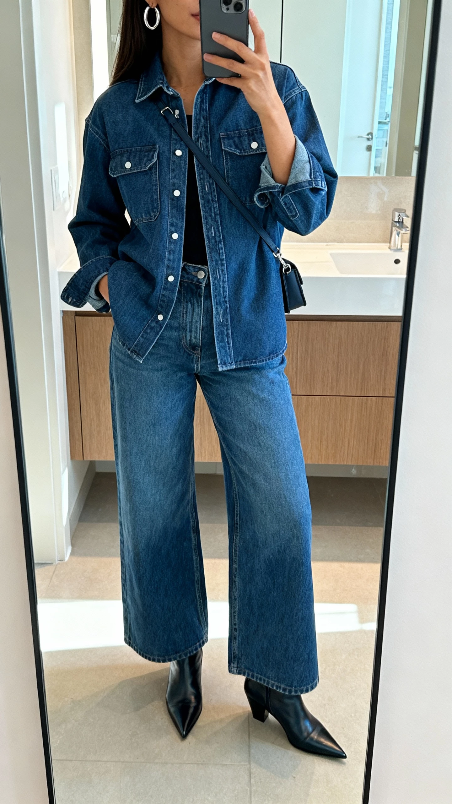 iPhone mirror selfie of a woman wearing wide leg denim jeans with a matching denim shirt/jacket (monochrome), pointed-toe boots, silver hoop earrings, minimalist crossbody, face not visible, bathroom mirror, bright natural light, natural iPhone photo quality.