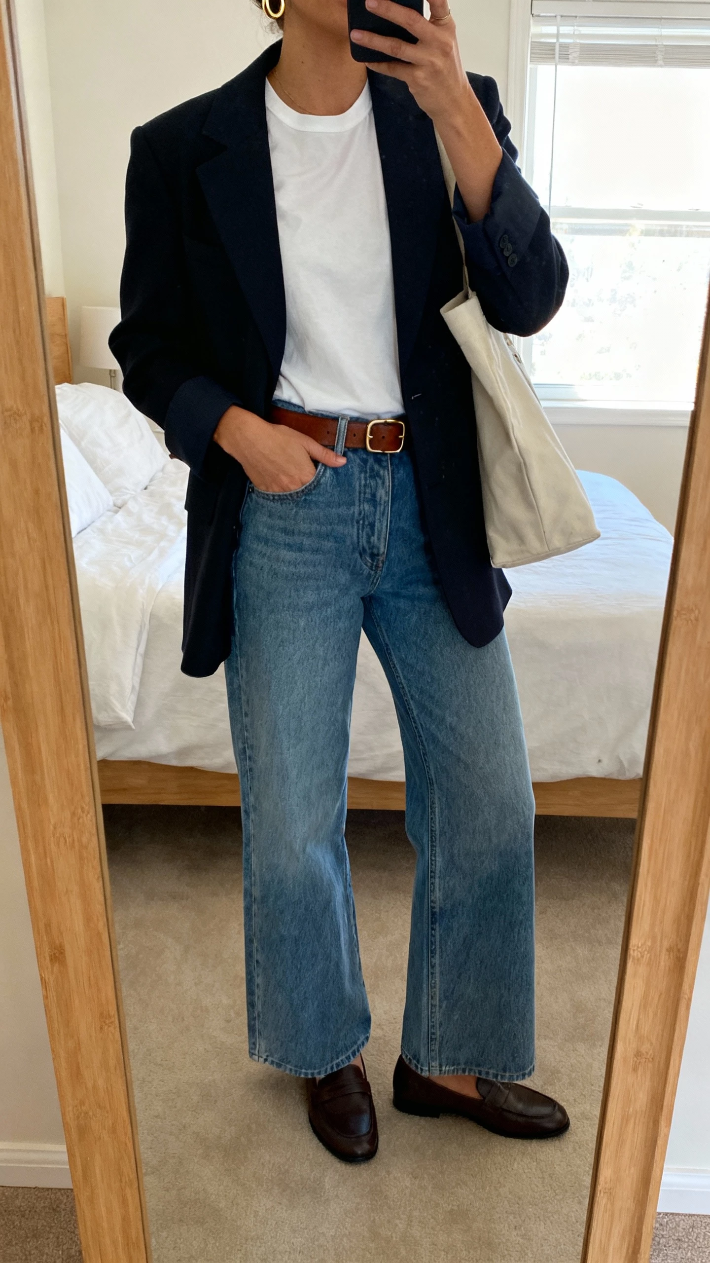 iPhone mirror selfie of a woman wearing wide leg jeans, crisp white tee, structured blazer, loafers, simple gold hoops, leather belt, tote bag, face not visible, cropped at neck in a bedroom mirror, soft window light, natural iPhone photo quality.
