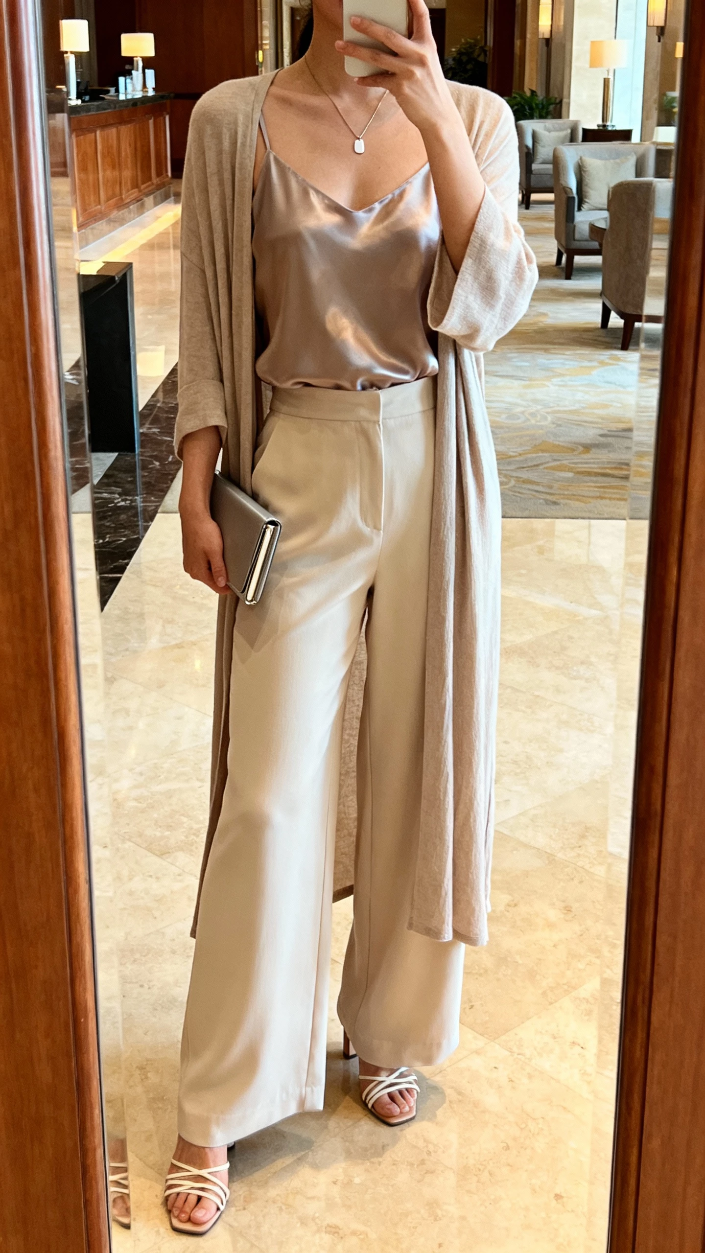iPhone mirror selfie of a woman wearing wide-leg trousers, a silk camisole, and a longline cardigan, strappy heels, minimalist pendant, slim clutch, face not visible, hotel lobby mirror, warm ambient lighting, natural iPhone photo quality.