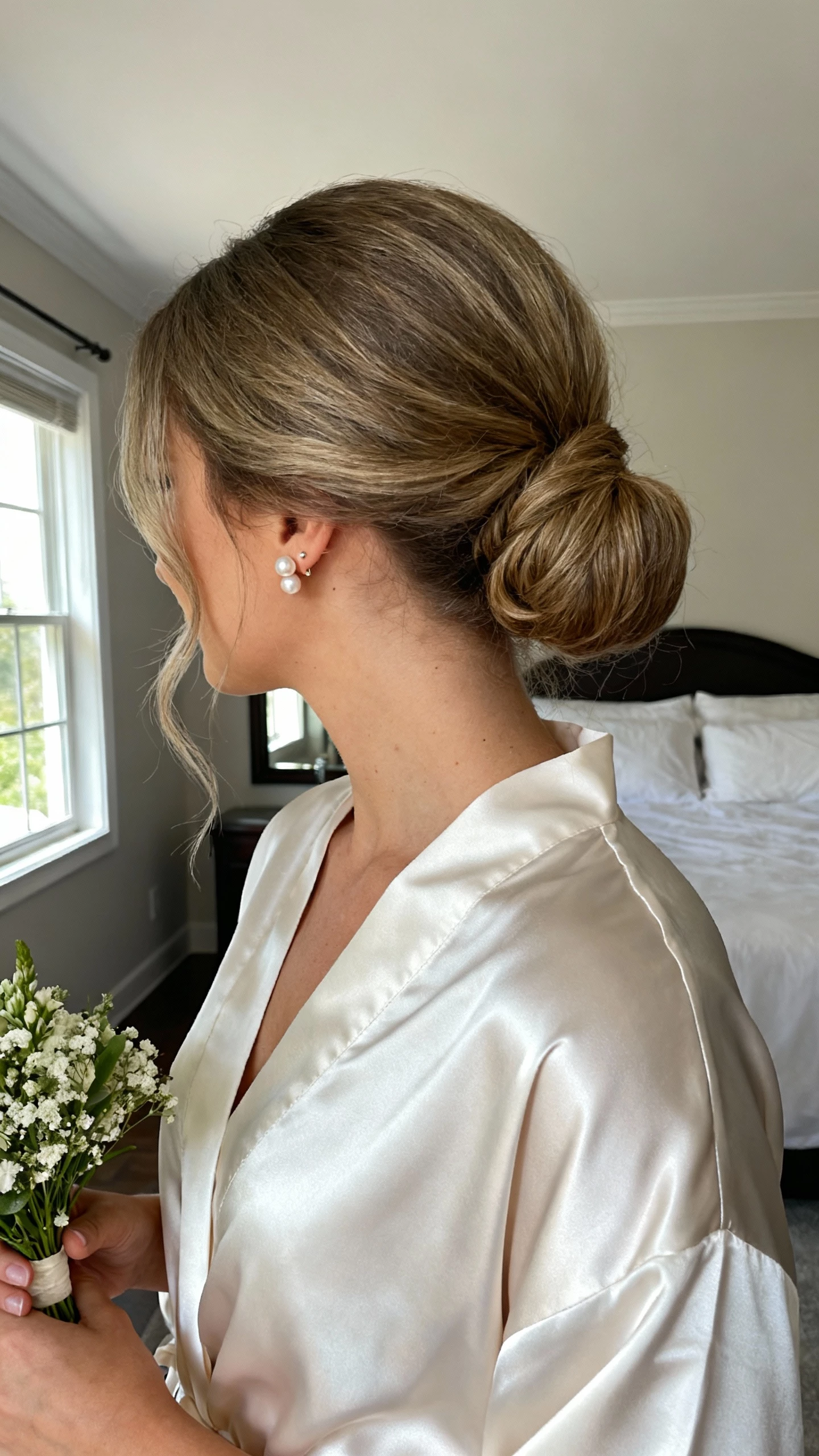 iPhone mirror selfie of a woman with a classic low chignon with a soft, airy finish, delicate pearl stud earrings, simple satin bridal robe, holding a small bouquet; face not visible, cropped at neck, bedroom mirror, natural window light, natural iPhone photo quality.