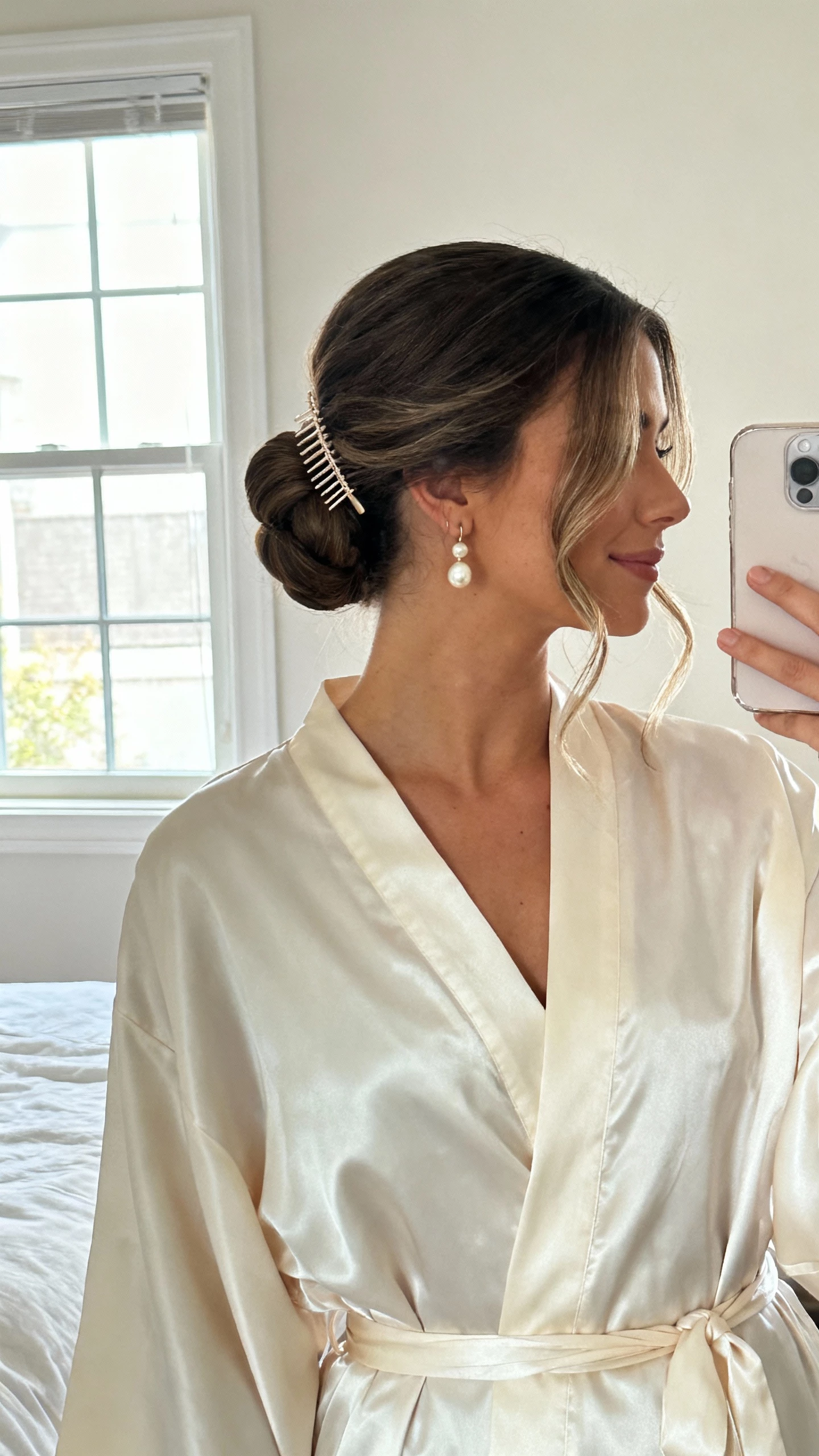 iPhone mirror selfie of a woman with a classic low chignon and soft face-framing tendrils, pearl drop earrings, satin wrap robe, delicate hair comb at the chignon, face not visible, cropped at neck, bedroom mirror with natural window light, natural iPhone photo quality.