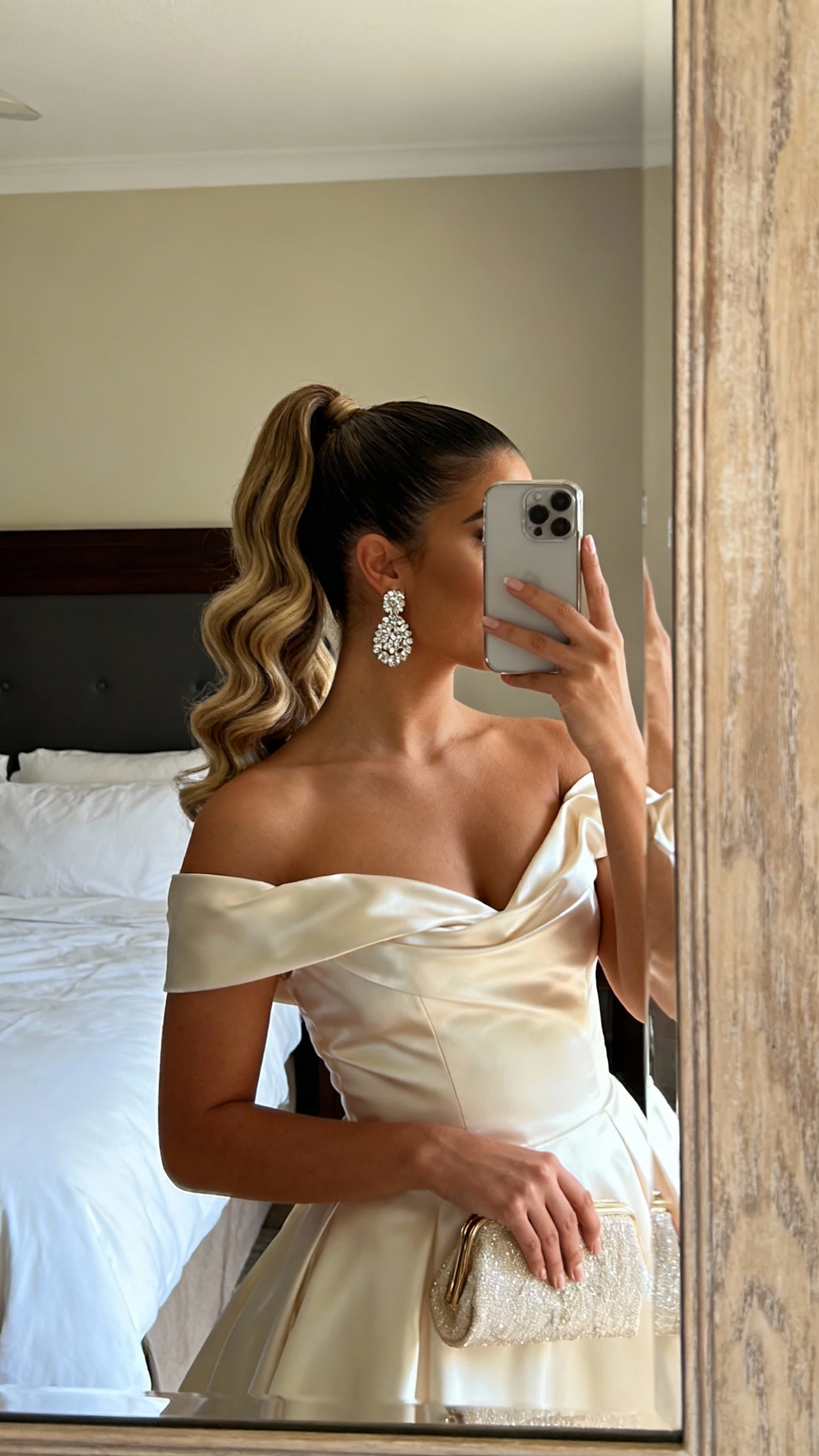 iPhone mirror selfie of a woman with a high ponytail styled in Hollywood waves, off-the-shoulder satin bridal gown, sparkly statement earrings, dainty clutch, face not visible, cropped at shoulders, bedroom mirror, warm indoor lighting, natural iPhone quality