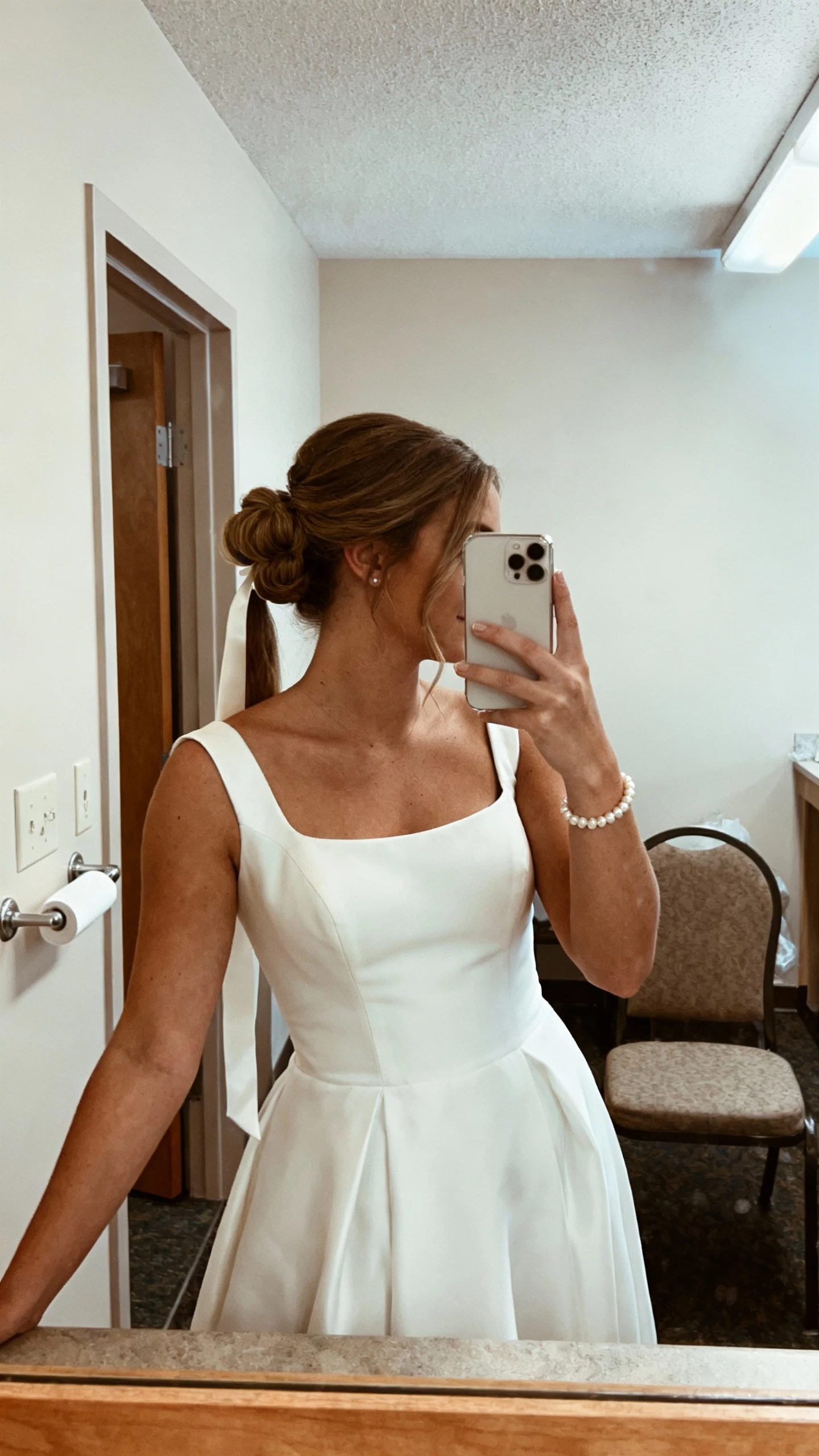 iPhone mirror selfie of a woman with a polished half-up bouffant tied with a ribbon tail, bateau-neck gown, pearl bracelet, face not visible, bridal suite mirror, diffused daylight, natural iPhone photo quality.