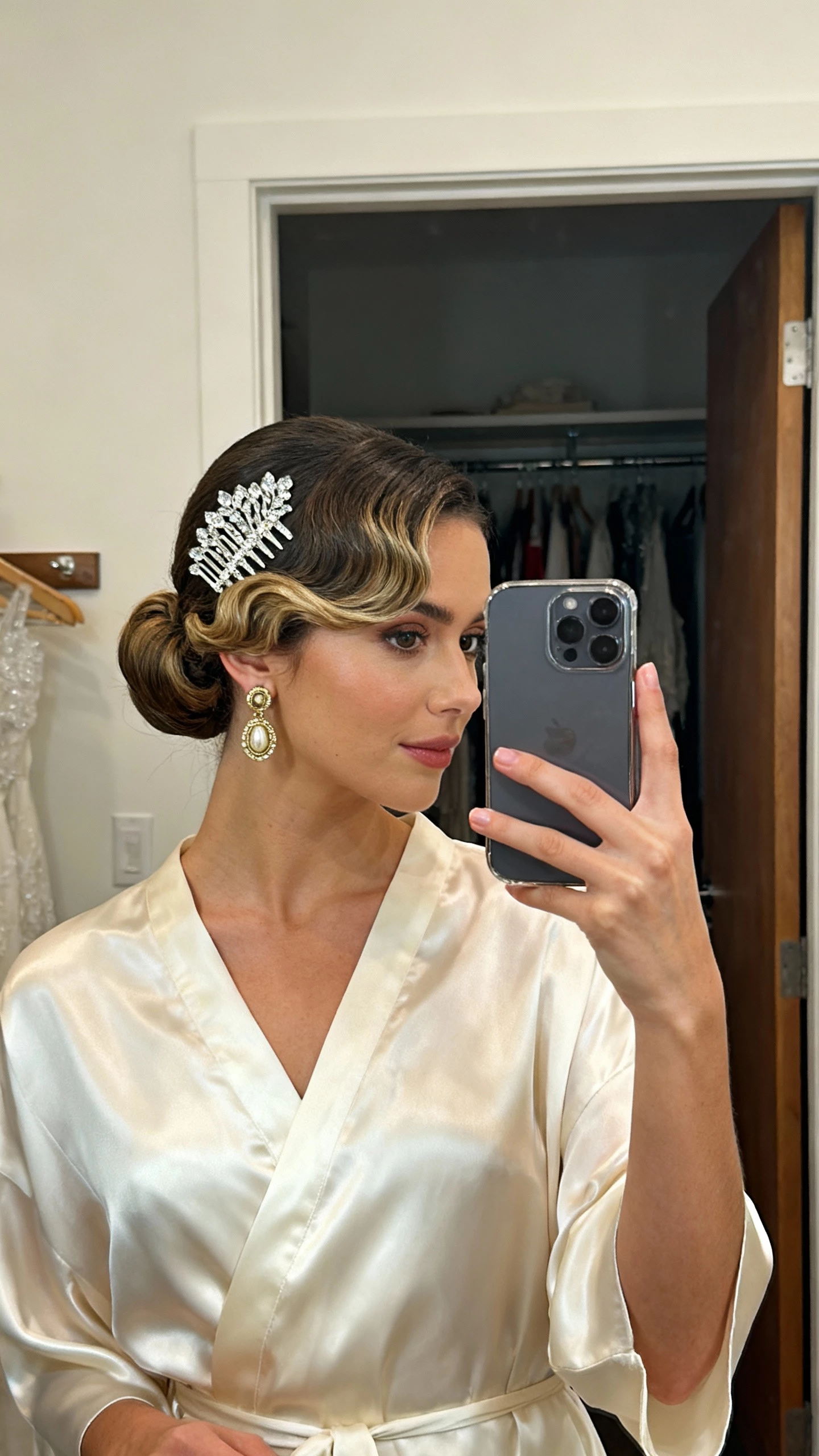 iPhone mirror selfie of a woman with a side-swept bun and vintage waves, wearing a silk bridal dress, statement crystal comb, and vintage-style earrings; face not visible, dressing room mirror, warm indoor lighting, casual iPhone shot.