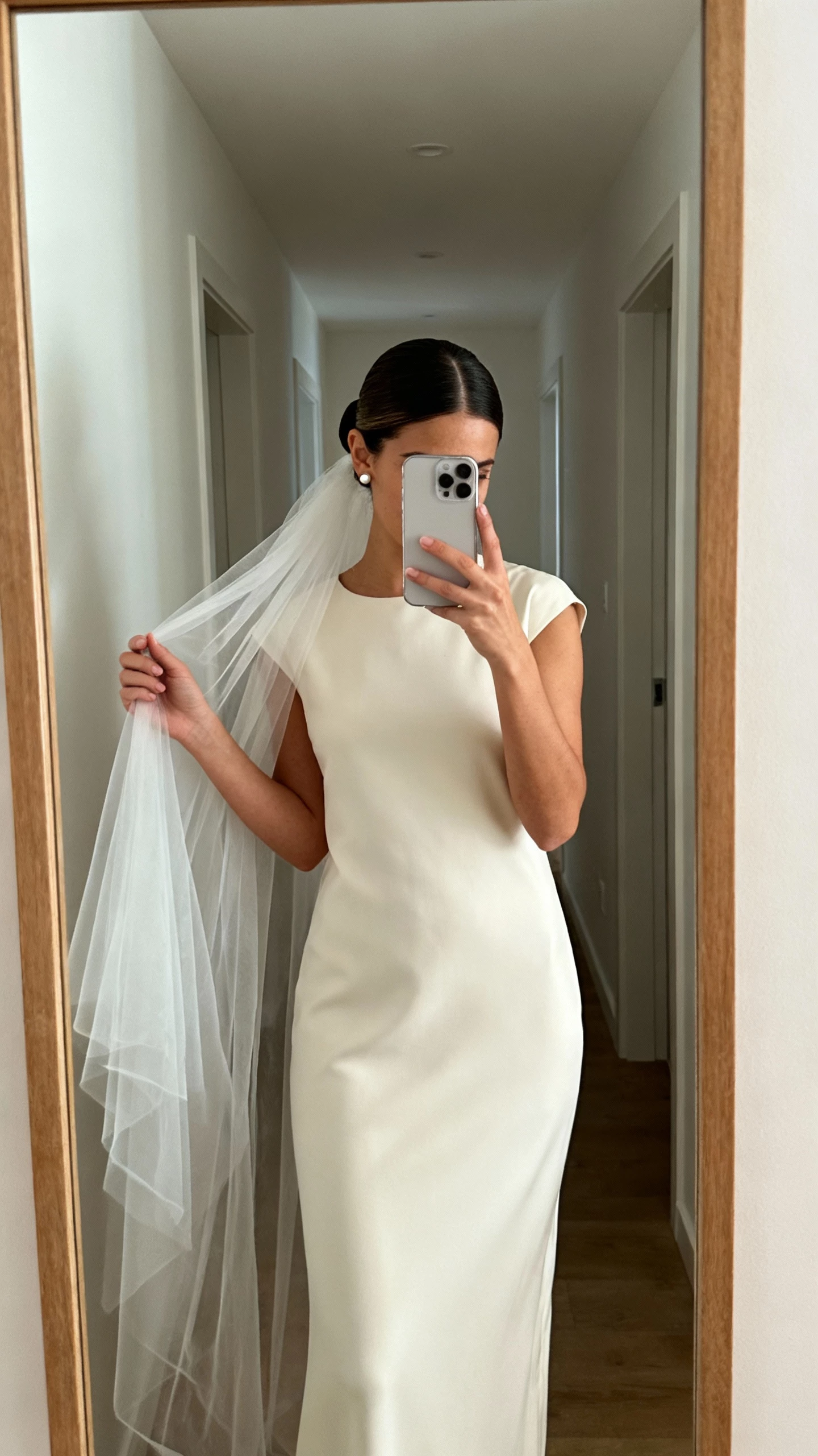 iPhone mirror selfie of a woman with a sleek center-part bun with a veil-ready base, veil draped over hand, minimalist column dress, stud earrings, face not visible, calm hallway mirror, natural indoor light, natural iPhone photo quality.