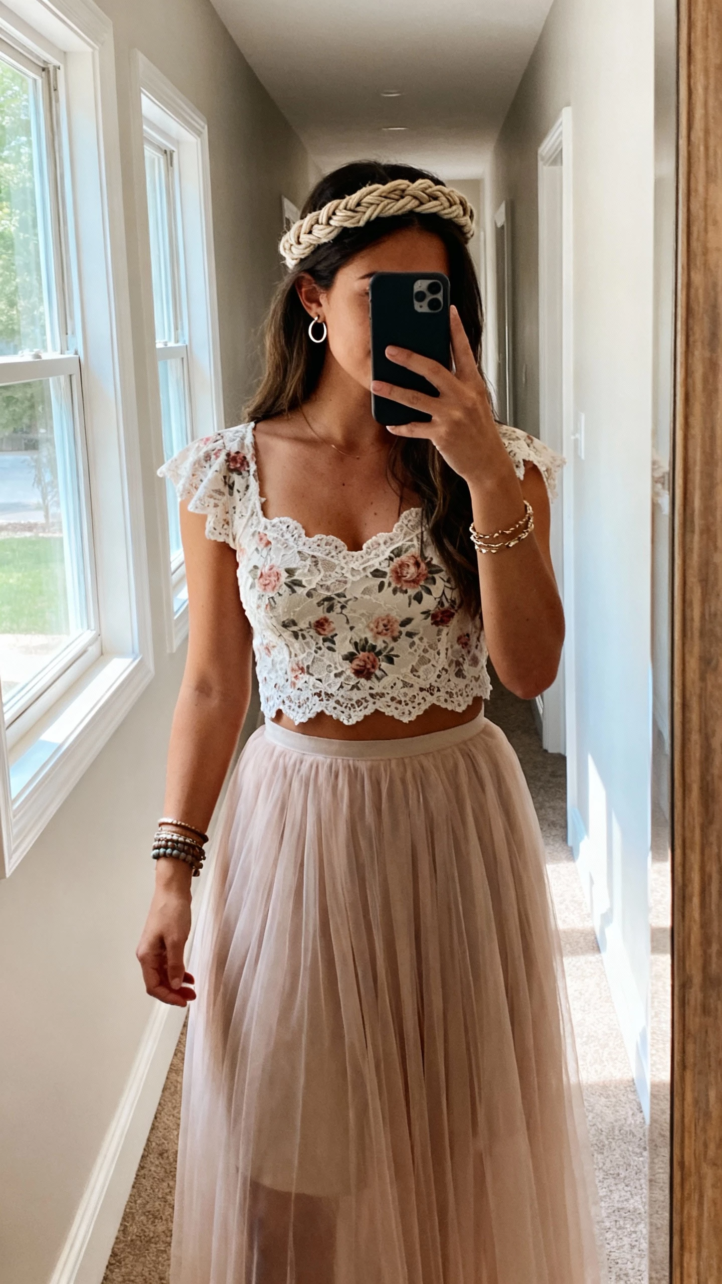 iPhone mirror selfie of a woman with a textured boho braid crown, wearing a flowy tulle skirt, floral lace top, layered bracelets, and small hoop earrings; face not visible, full-length hallway mirror, afternoon window light, natural iPhone photo quality.