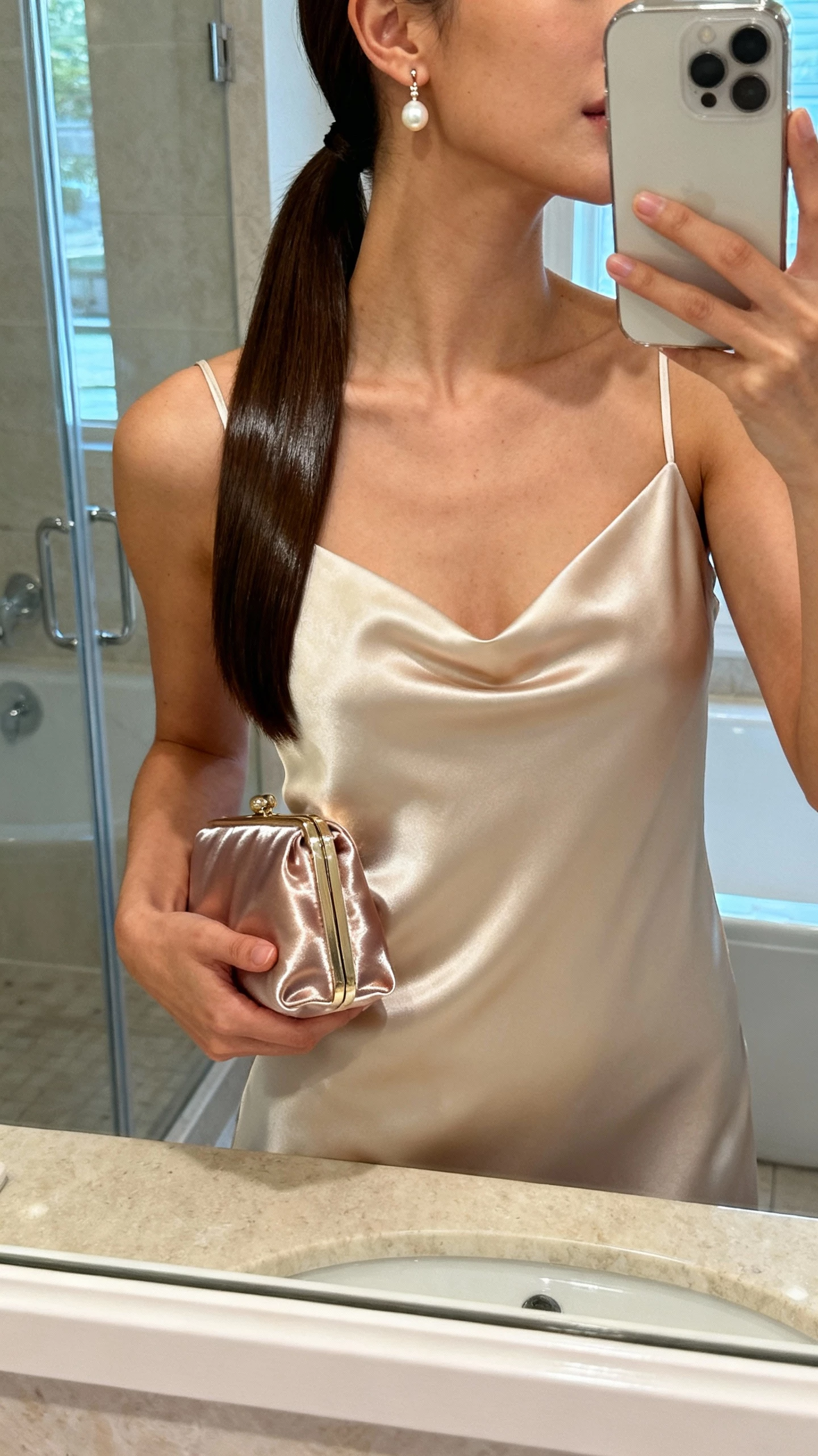 iPhone mirror selfie of a woman with a velvet-gloss sleek low ponytail, minimalist satin bridal slip dress, slim pearl drop earrings, glossy clutch, face not visible, cropped at neck, bathroom mirror with soft indoor light, natural iPhone photo quality
