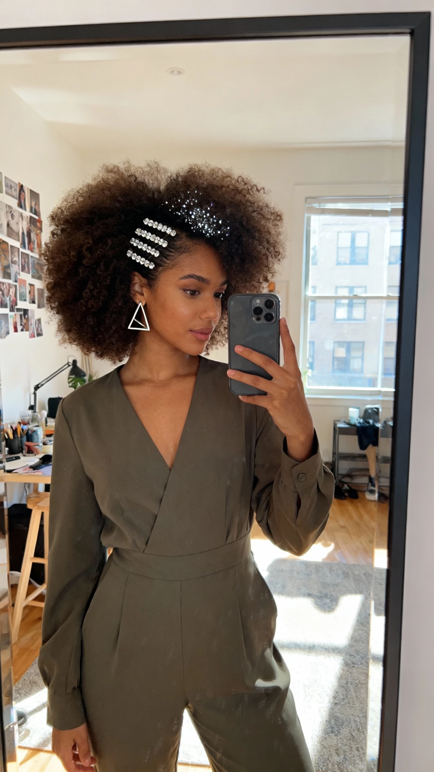 iPhone mirror selfie of a woman with a voluminous curly afro and a sparkling part, structured modern jumpsuit, crystal hair pins along the part, geometric earrings, face not visible, studio apartment mirror, bright daylight, natural iPhone photo.