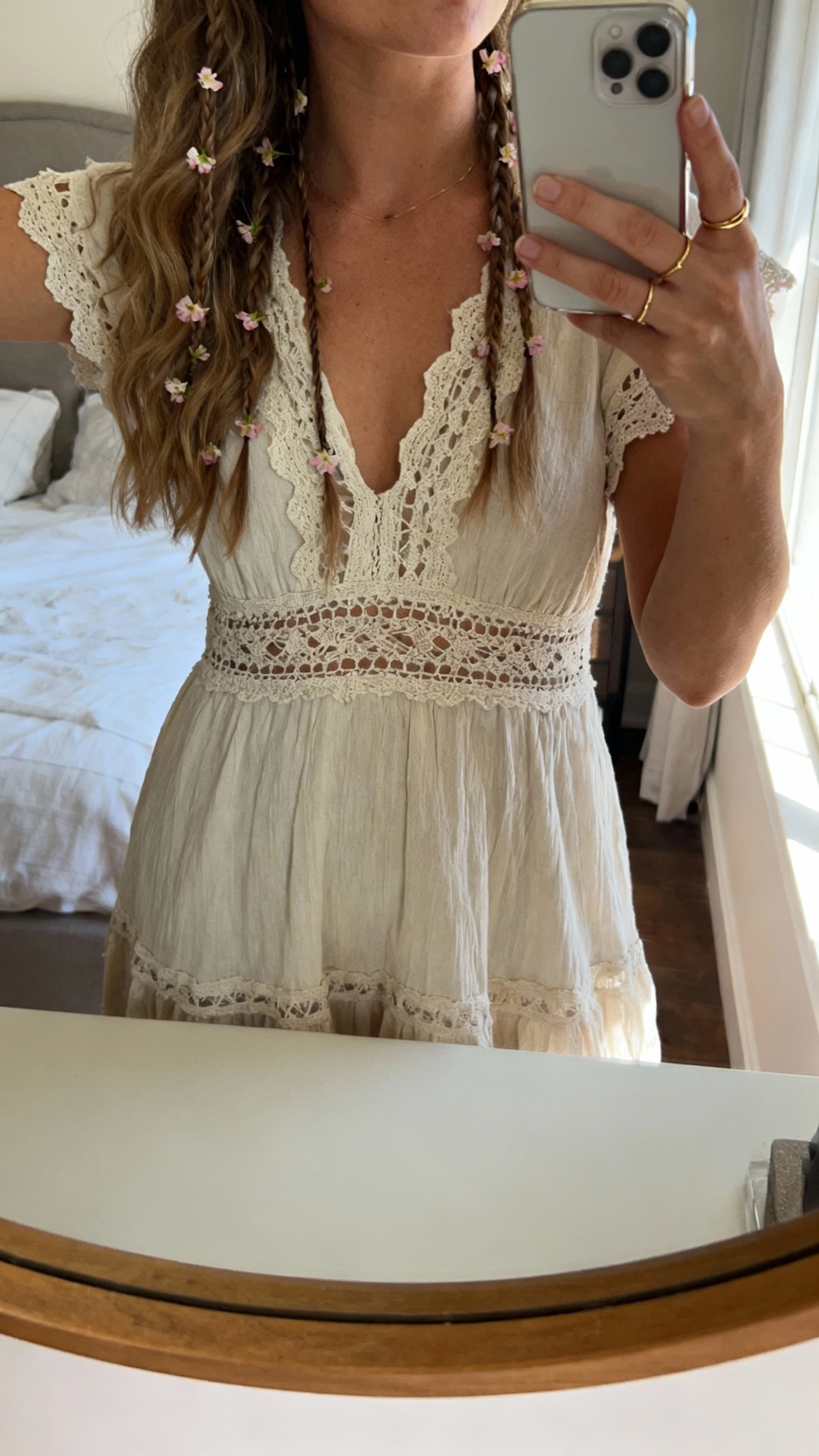 iPhone mirror selfie of a woman with barely-there waves, thin baby braids threaded with tiny petals, wearing a simple boho lace dress and delicate gold rings, face not visible, cropped at neck, bedroom mirror with morning window light, natural iPhone photo quality.