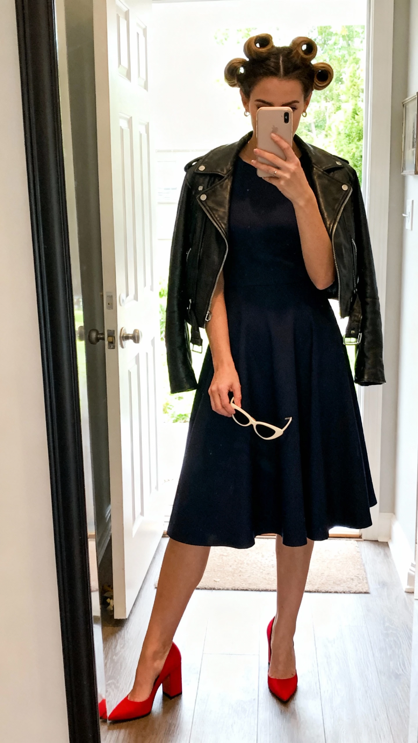 iPhone mirror selfie of a woman with victory rolls and a modern edge, tea-length fit-and-flare dress, leather moto jacket draped over shoulders, red block-heel pumps, cat-eye sunglasses in hand; face not visible, entryway mirror, daylight from door, natural iPhone photo.