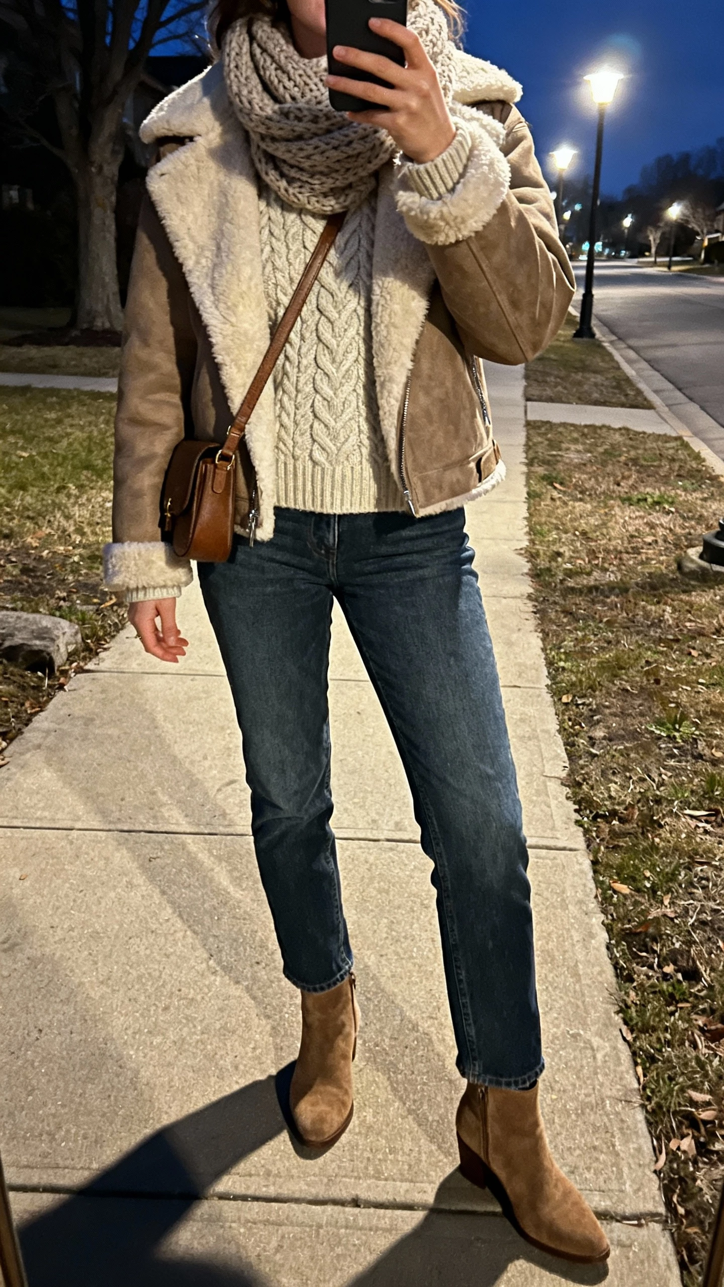 iPhone night sidewalk selfie of a woman wearing a shearling-lined jacket over a cable-knit sweater, straight jeans, suede ankle boots, knit scarf, crossbody bag, face not visible, streetlights and cool evening glow, natural iPhone photo quality