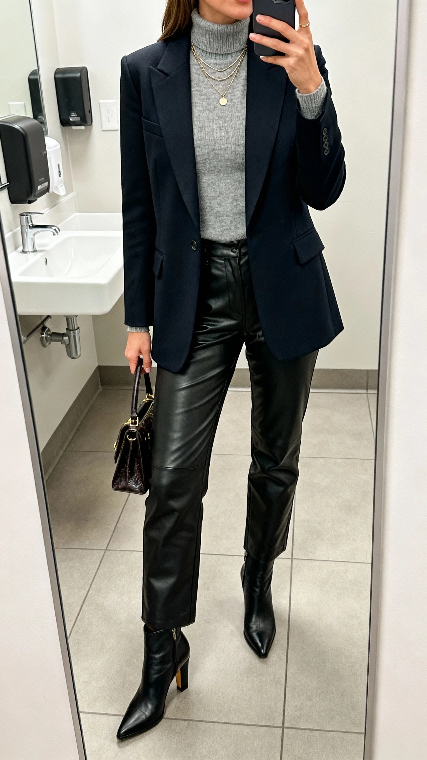 iPhone office bathroom mirror selfie of a woman in a fitted cashmere-blend turtleneck, tailored single-breasted blazer, faux leather straight-leg trousers, heeled ankle boots, delicate layered necklaces, and a classic handbag, face not visible, cropped at shoulders, bright indoor lighting, natural iPhone aesthetic.