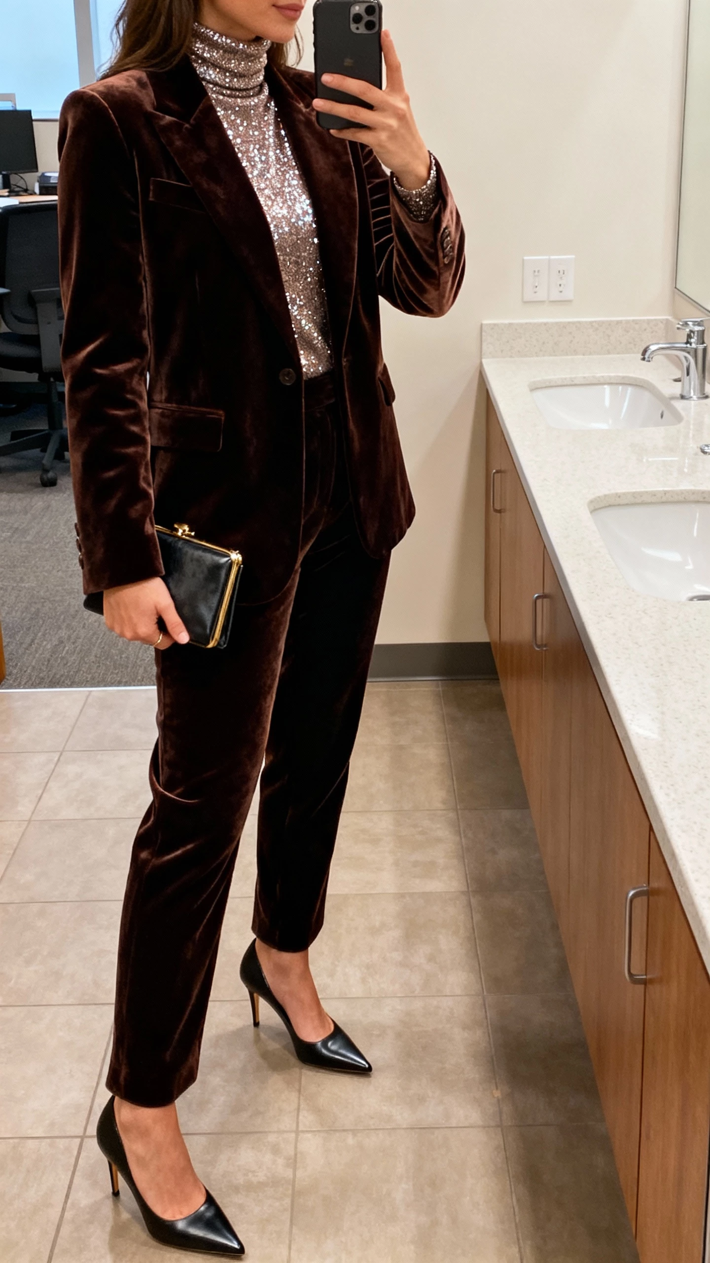 iPhone office-bathroom mirror selfie of a woman in a velvet suit with a sparkly turtleneck, pointed heels, and a small clutch, face not visible, side angle, warm indoor lighting, natural iPhone photo quality.