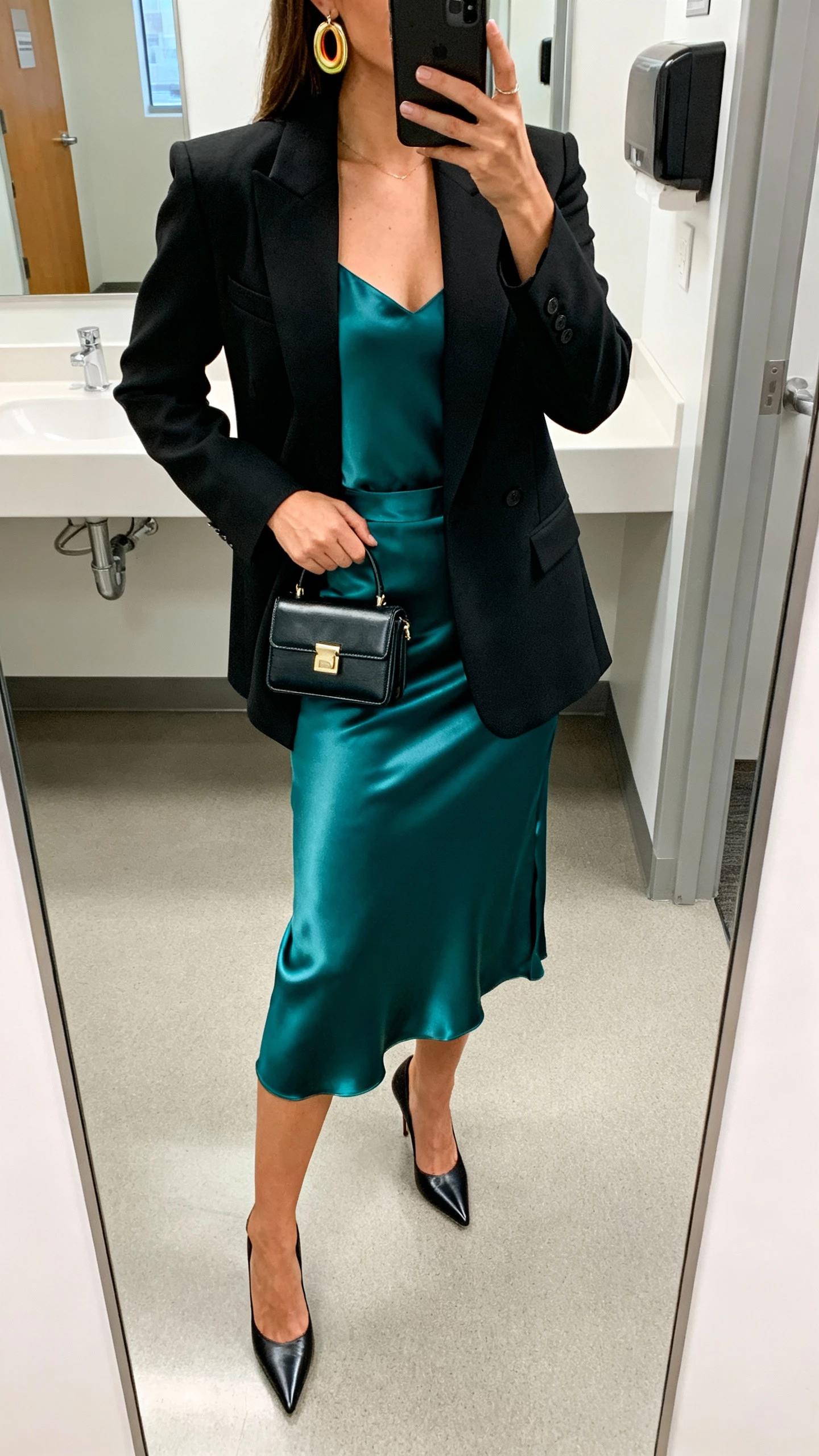 iPhone office-bathroom mirror selfie of a woman wearing a sharp power blazer over a satin midi skirt set, sleek camisole, pointed heels, bold earrings, and a structured mini bag—face not visible, full-length mirror, bright indoor lighting, natural iPhone photo quality.