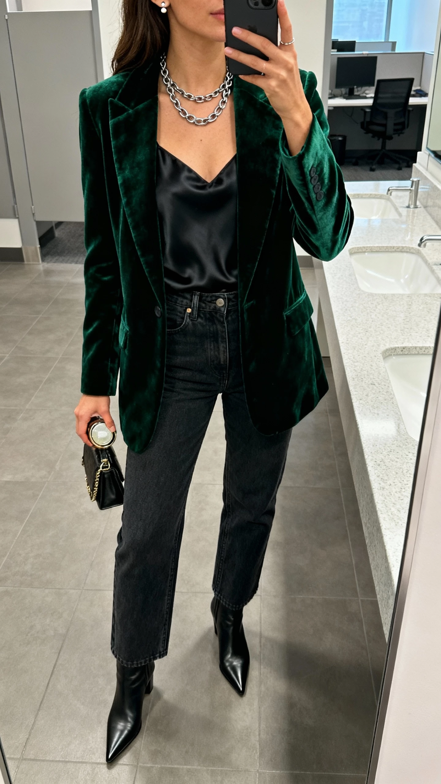 iPhone office bathroom mirror selfie of a woman wearing a deep green velvet blazer over a black silk camisole, high-rise straight-leg dark jeans, pointed-toe ankle boots, layered chain necklaces and small stud earrings, holding a compact bag; face not visible, indoor lighting, natural iPhone photo quality.