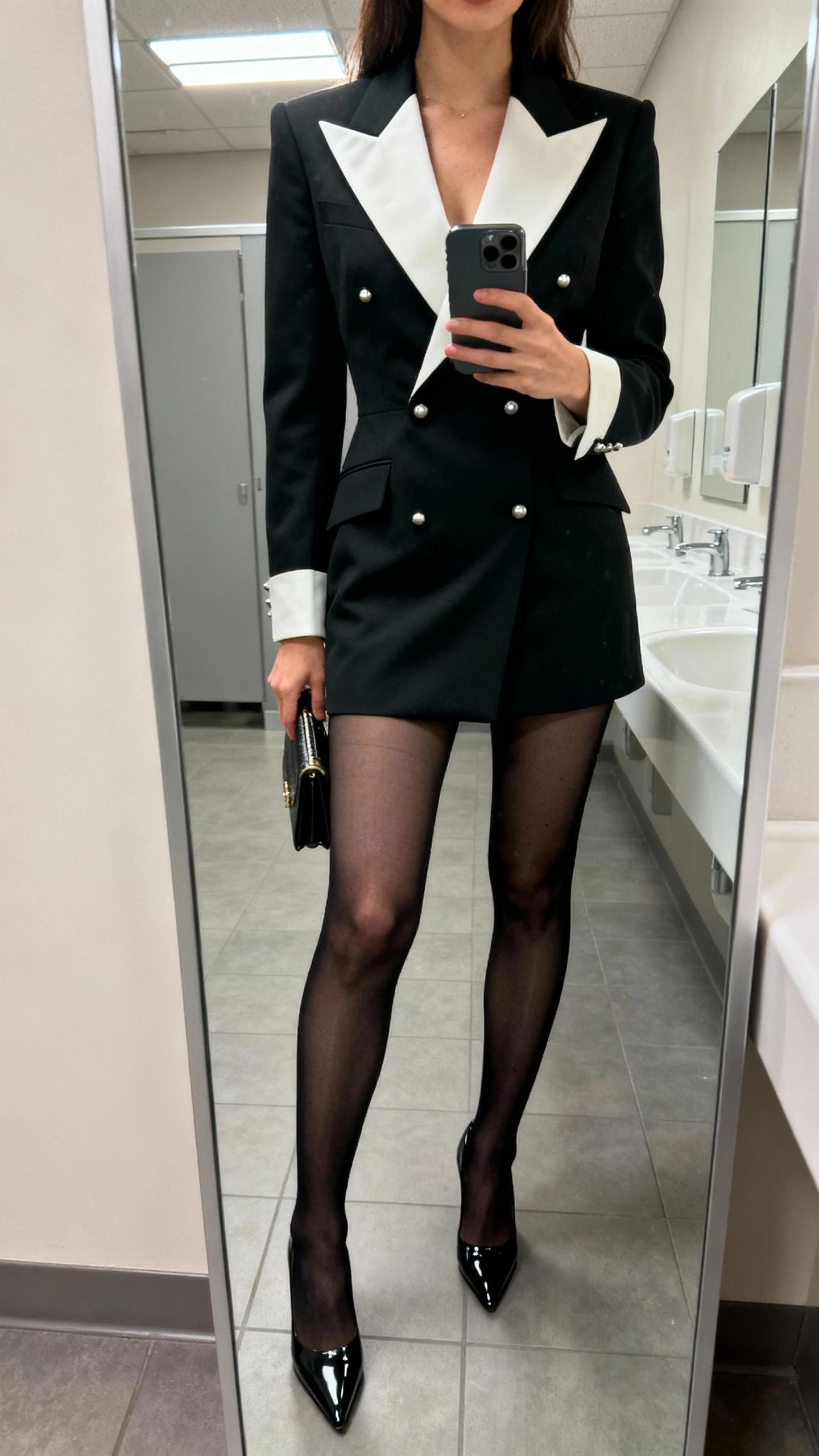 iPhone office-bathroom mirror selfie of a woman wearing a tailored tuxedo dress with peak lapels, sheer black tights, pointed patent pumps, minimalist studs, small clutch, face not visible, cropped at shoulders, overhead fluorescent light, natural iPhone aesthetic.