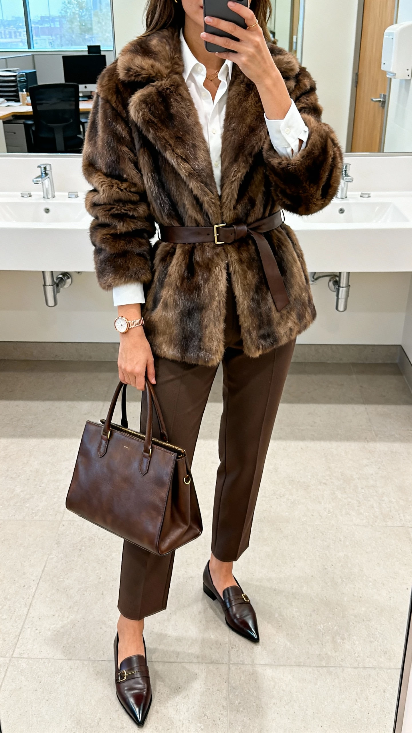 iPhone office bathroom mirror selfie of a woman wearing a belted fur jacket over tailored trousers and a crisp blouse, pointed-toe loafers, structured leather tote, delicate watch, face not visible, bright office lighting, natural iPhone photo quality.