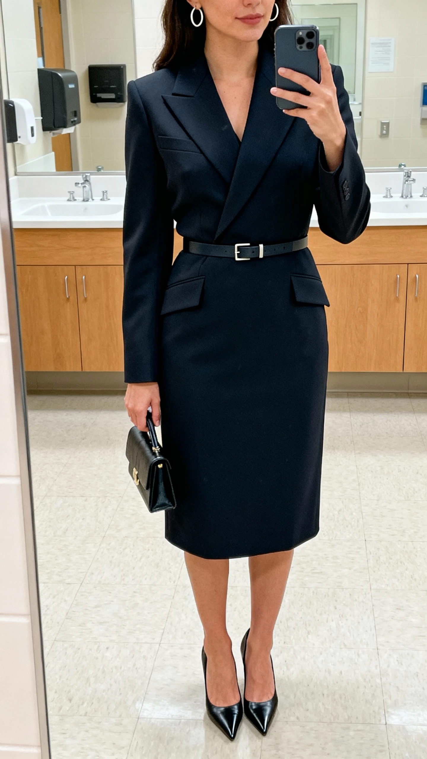 iPhone office-bathroom mirror selfie of a woman wearing a chic courthouse suit-dress (tailored blazer dress) with sharp lapels, minimalist belt, pointed-toe heels, small structured clutch, and simple hoop earrings, face not visible, bright indoor lighting, natural iPhone quality.