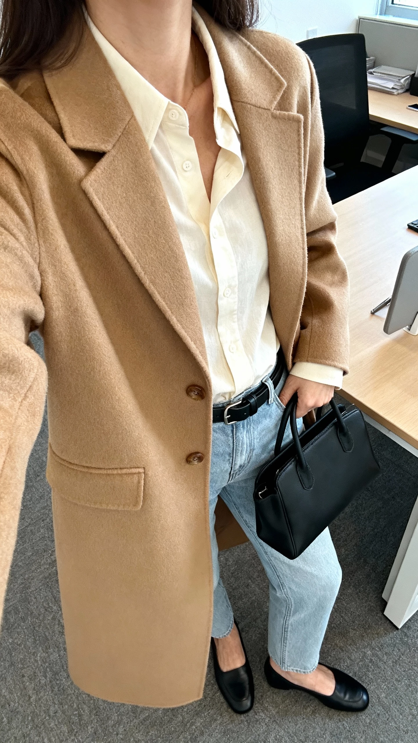 iPhone office desk selfie cropped at neck showing a woman in a soft cream blouse tucked into light blue jeans, camel tailored coat, minimalist black belt, black loafers, structured black tote, face not visible, morning office light, natural iPhone photo quality