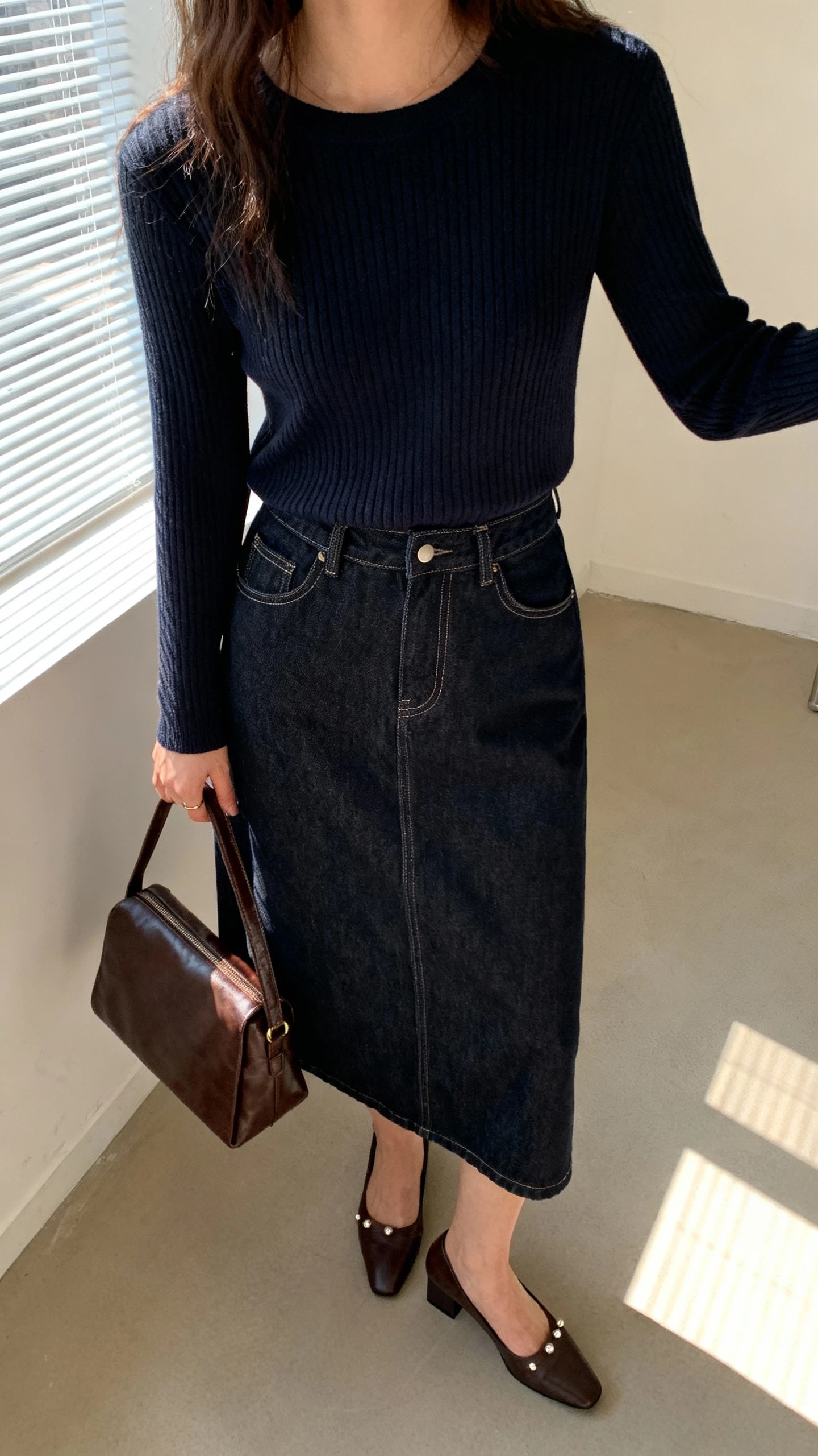 iPhone office mirror selfie of a woman in a dark denim midi skirt with a slim knit sweater tucked in, low block-heel pumps, simple studs, leather satchel, face not visible, cropped at neck, indoor overhead plus window light, natural iPhone photo quality