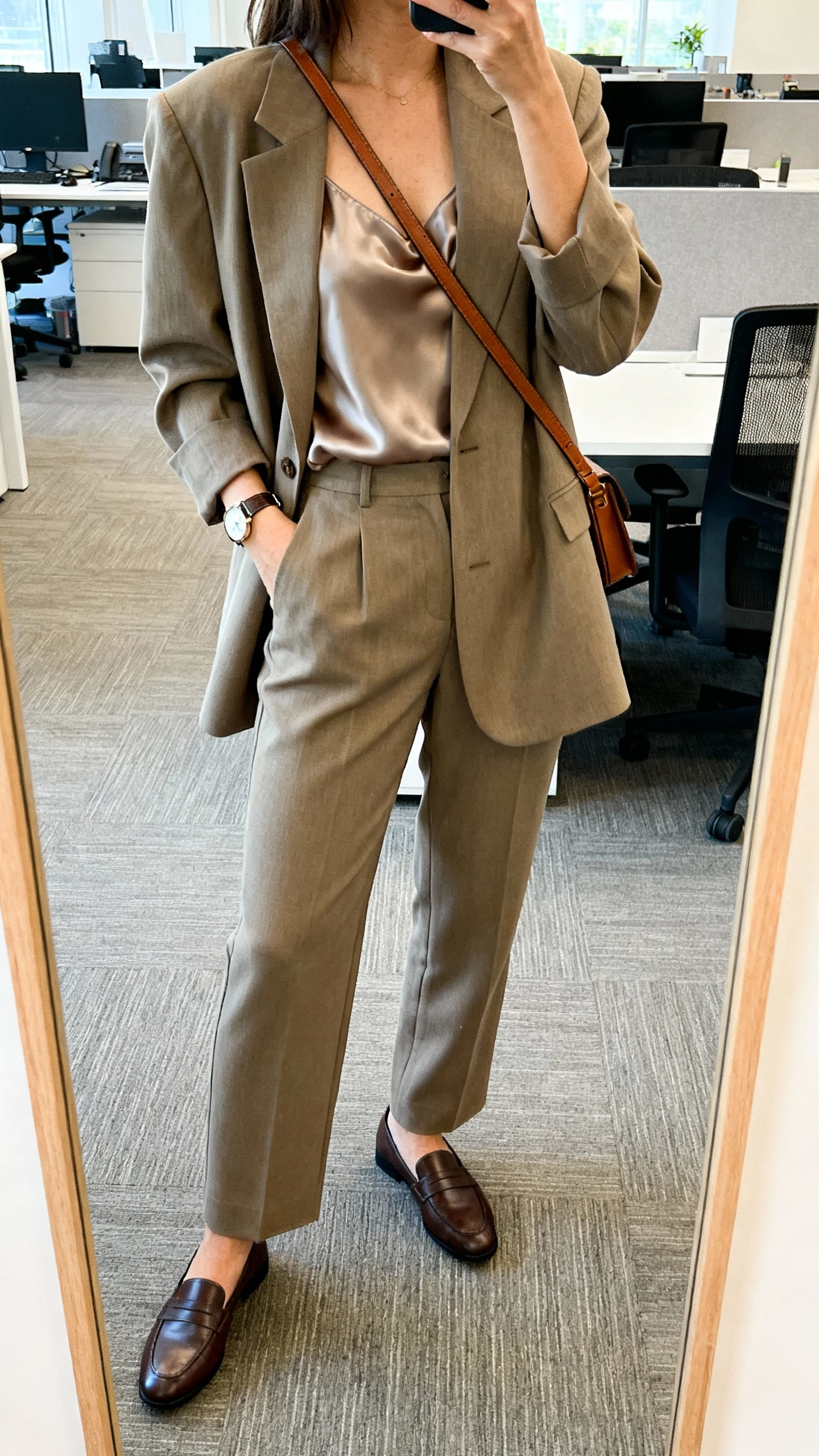 iPhone office mirror selfie of a woman in a relaxed suit set (boxy blazer and straight trousers) with a silk camisole, loafers, slim leather crossbody, watch, face not visible, cropped at shoulders, bright office light, natural iPhone photo quality.