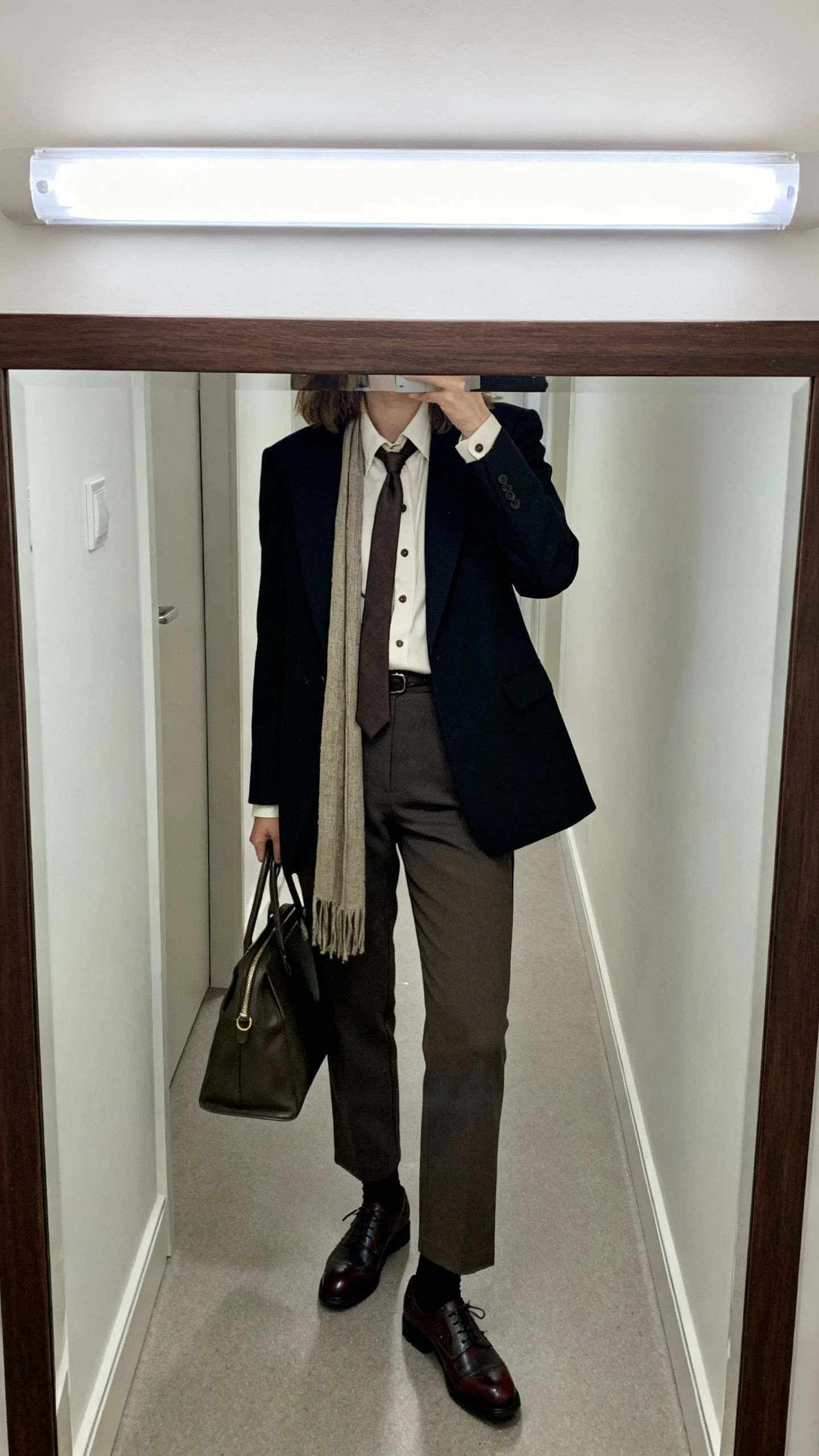 iPhone office mirror selfie of a woman in menswear-inspired layers: sharp blazer over a crisp button-up, long scarf tied like a tie knot, tailored trousers, brogues, and a structured tote, face not visible, cropped at shoulders, fluorescent-lit hallway mirror, natural iPhone aesthetic.