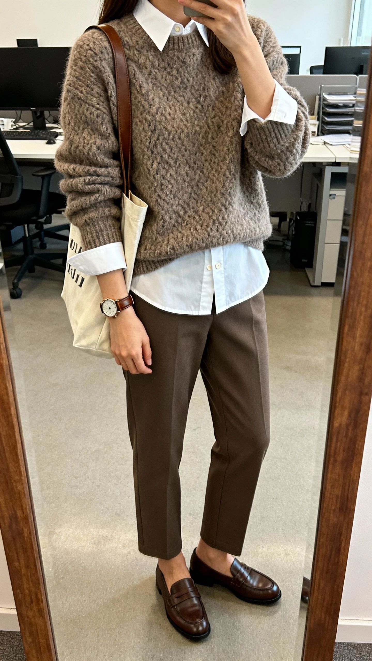 iPhone office-mirror selfie of a woman layering a chunky crewneck sweater over a crisp white button-down (collar and hem peeking), straight-leg trousers, loafers, leather watch, tote bag, face not visible, cropped at shoulders, morning office light, natural iPhone photo quality.