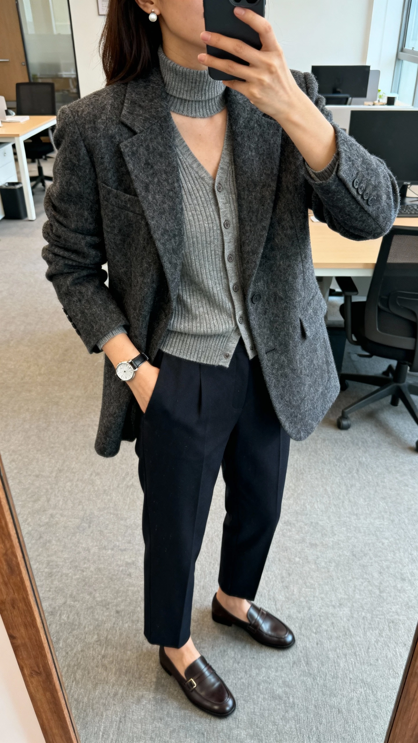 iPhone office mirror selfie of a woman wearing a charcoal wool blazer layered over a fine-knit turtleneck and a thin cardigan, tailored dark trousers, loafers, watch and stud earrings, face not visible, cropped at shoulders, natural office daylight, iPhone photo quality.