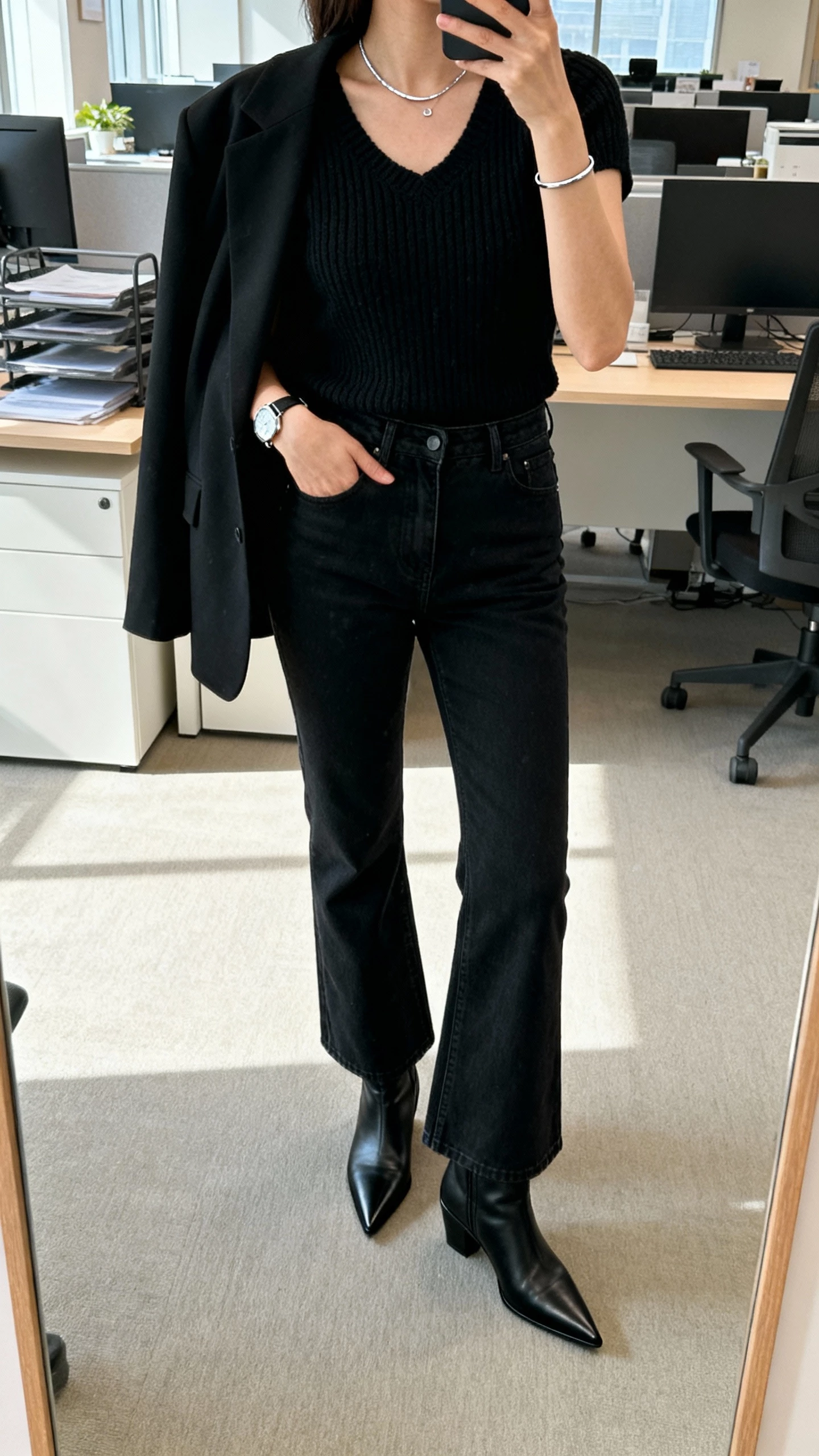 iPhone office mirror selfie of a woman wearing a monochrome set: black knit top, black bootcut jeans, black pointed boots, black blazer draped over shoulders, minimalist silver jewelry, slim watch; face not visible, cropped at neck, bright morning office light, natural iPhone photo quality.