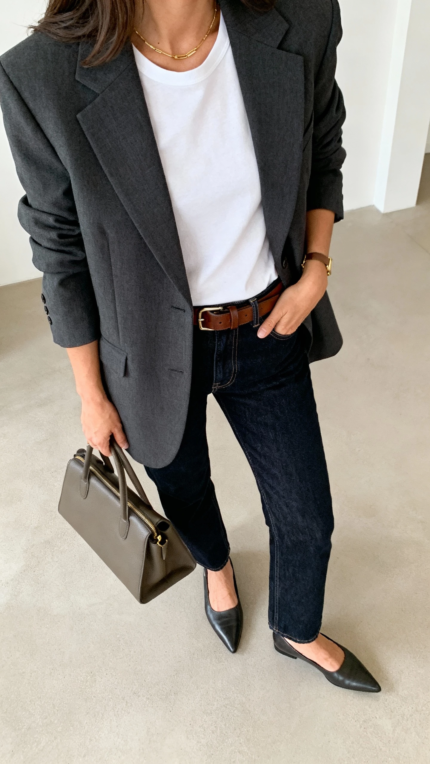 iPhone office mirror selfie of a woman wearing a tailored charcoal blazer over a white tee with dark-wash straight-leg denim, pointed loafers, leather belt, structured tote, minimal gold jewelry, face not visible, side angle, indoor overhead lighting, natural iPhone photo quality.