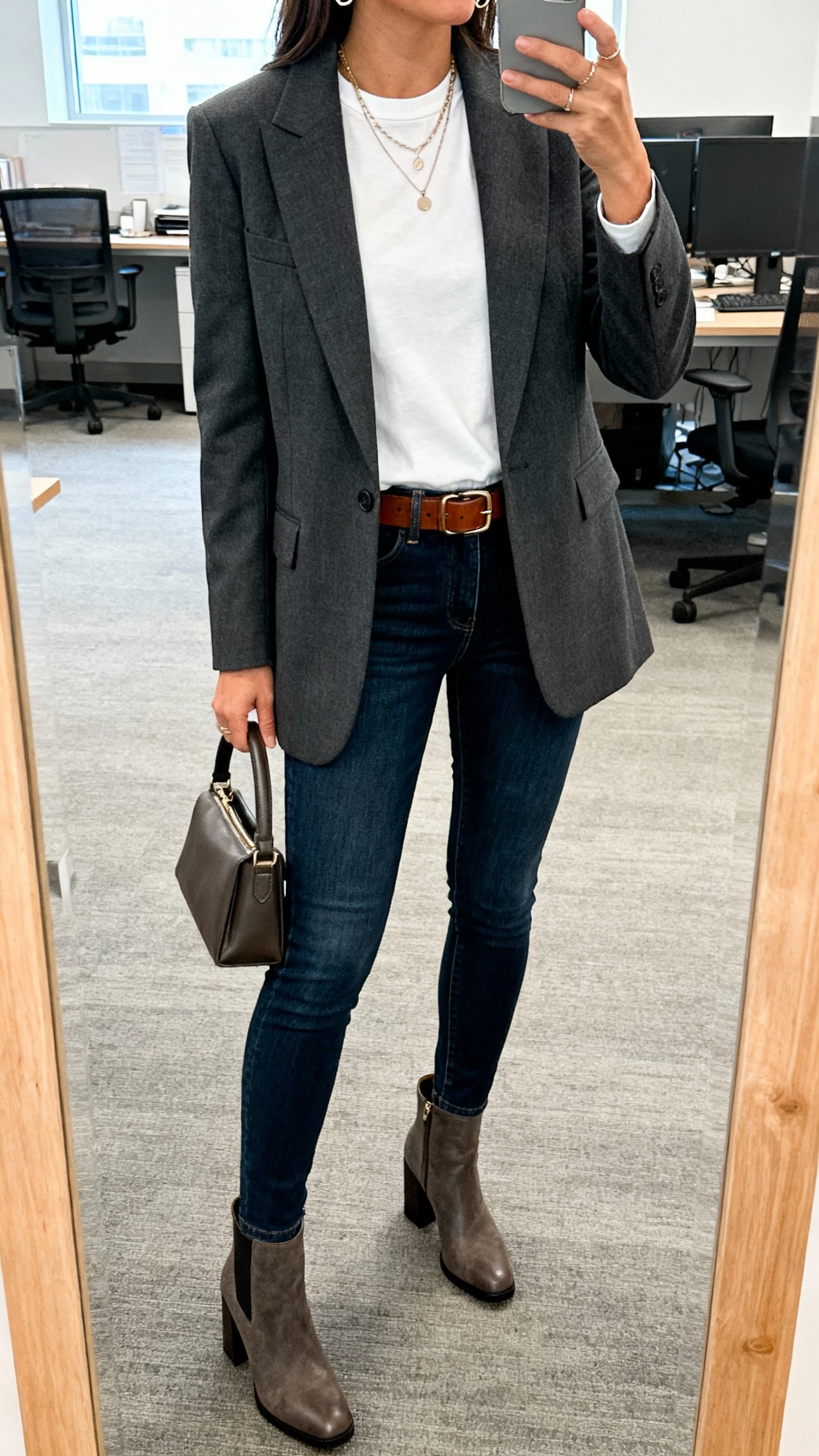 iPhone office mirror selfie of a woman wearing a tailored charcoal blazer over a white crewneck tee, dark blue slim jeans, block-heel ankle boots, leather belt, layered necklaces, small hoops, and a top-handle bag; cropped at neck, indoor lighting, face not visible, casual iPhone photo.