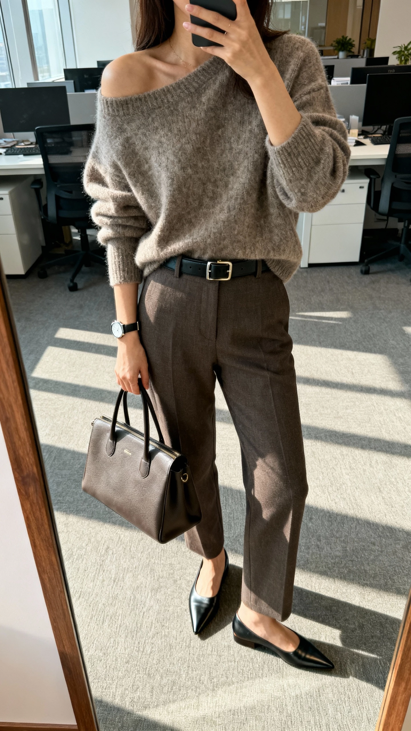 iPhone office mirror selfie of a woman wearing an oversized sweater tucked into tailored trousers, sleek belt, pointed loafers, minimalist watch, and structured tote, face not visible, shoulder-down crop, bright morning office light, natural iPhone photo quality.