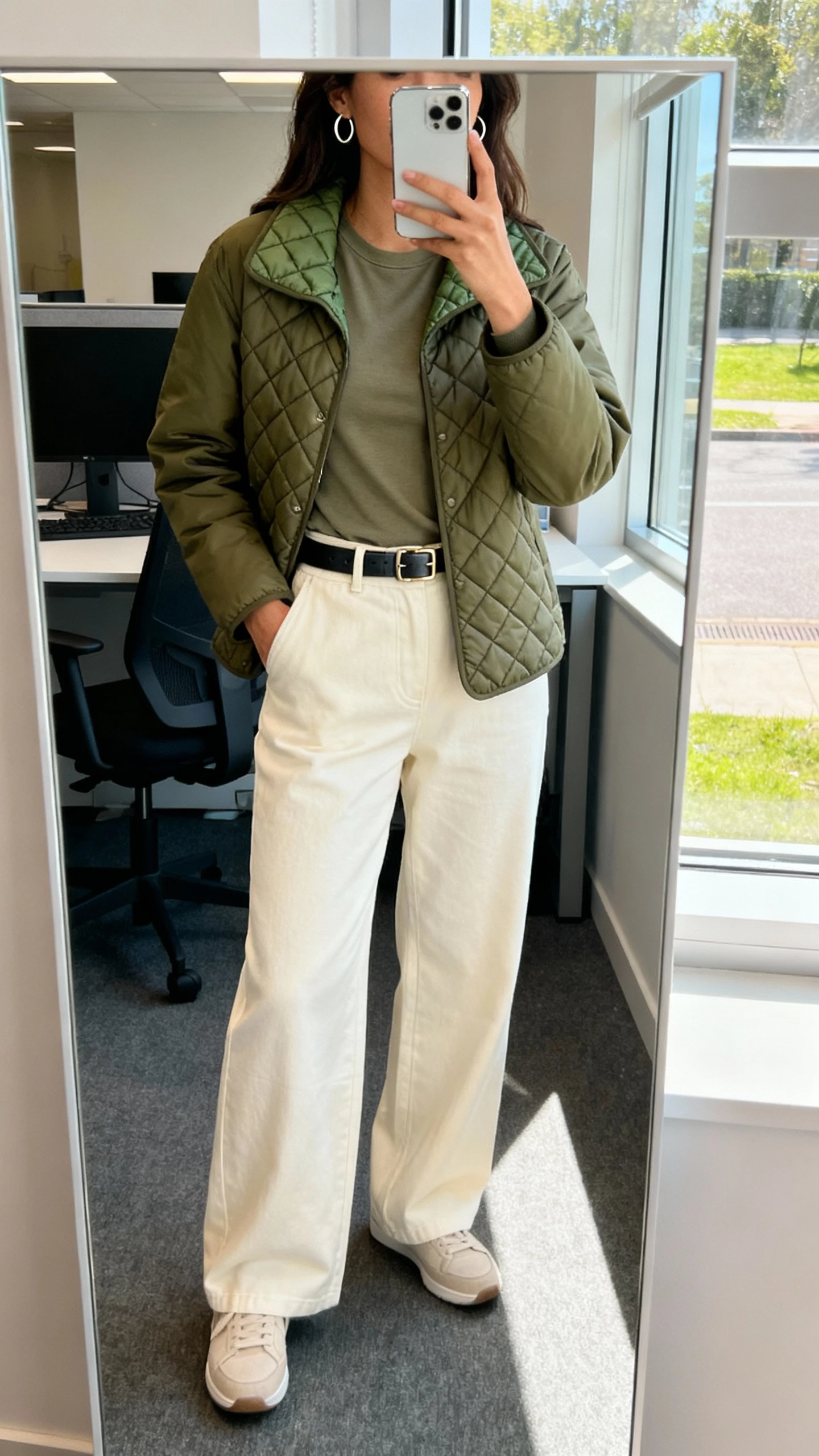 iPhone office mirror selfie of a woman wearing an olive quilted liner jacket with cream wide-leg pants, fitted tee, minimalist sneakers, sleek belt, simple hoops, face not visible, bright indoor office light by window, natural iPhone photo quality
