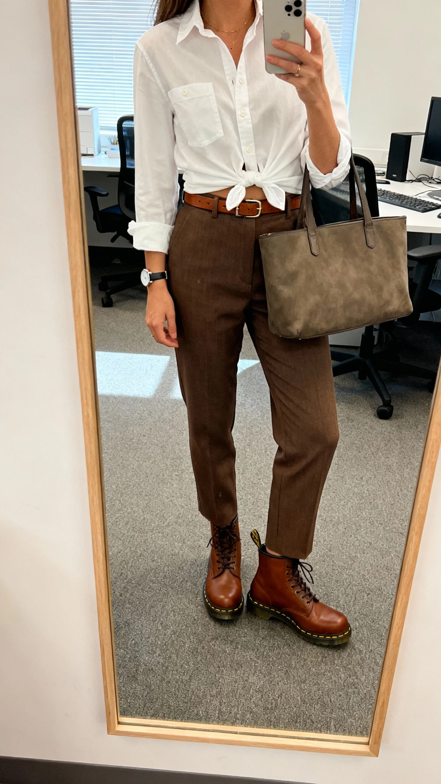 iPhone office mirror selfie of a woman wearing brown Doc Martens with tailored high-waist trousers, crisp white button-down half-tucked, slim leather belt, minimal watch, and structured tote, face not visible, cropped at shoulders, morning office light, natural iPhone quality.