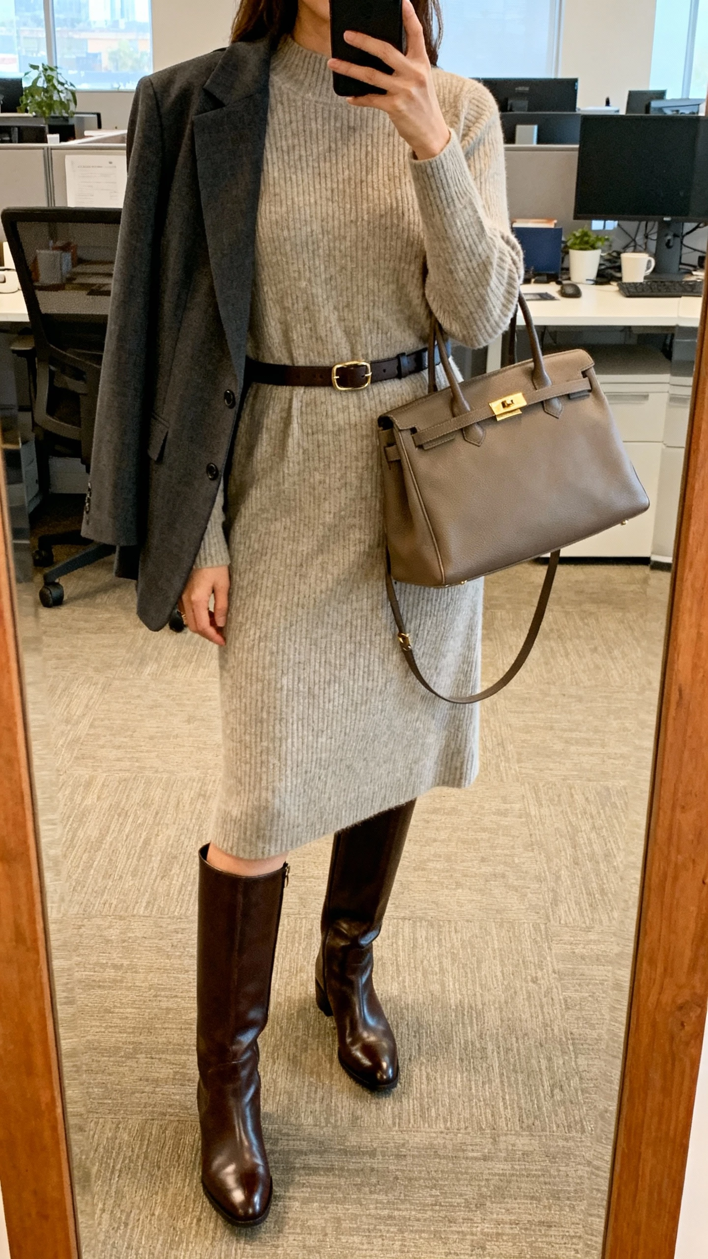 iPhone office mirror selfie of a woman wearing sleek knee-high leather boots with a tailored midi sweater dress, structured blazer draped over shoulders, slim belt, and tote bag, face not visible, cropped at shoulders, warm office lighting, natural iPhone photo quality.