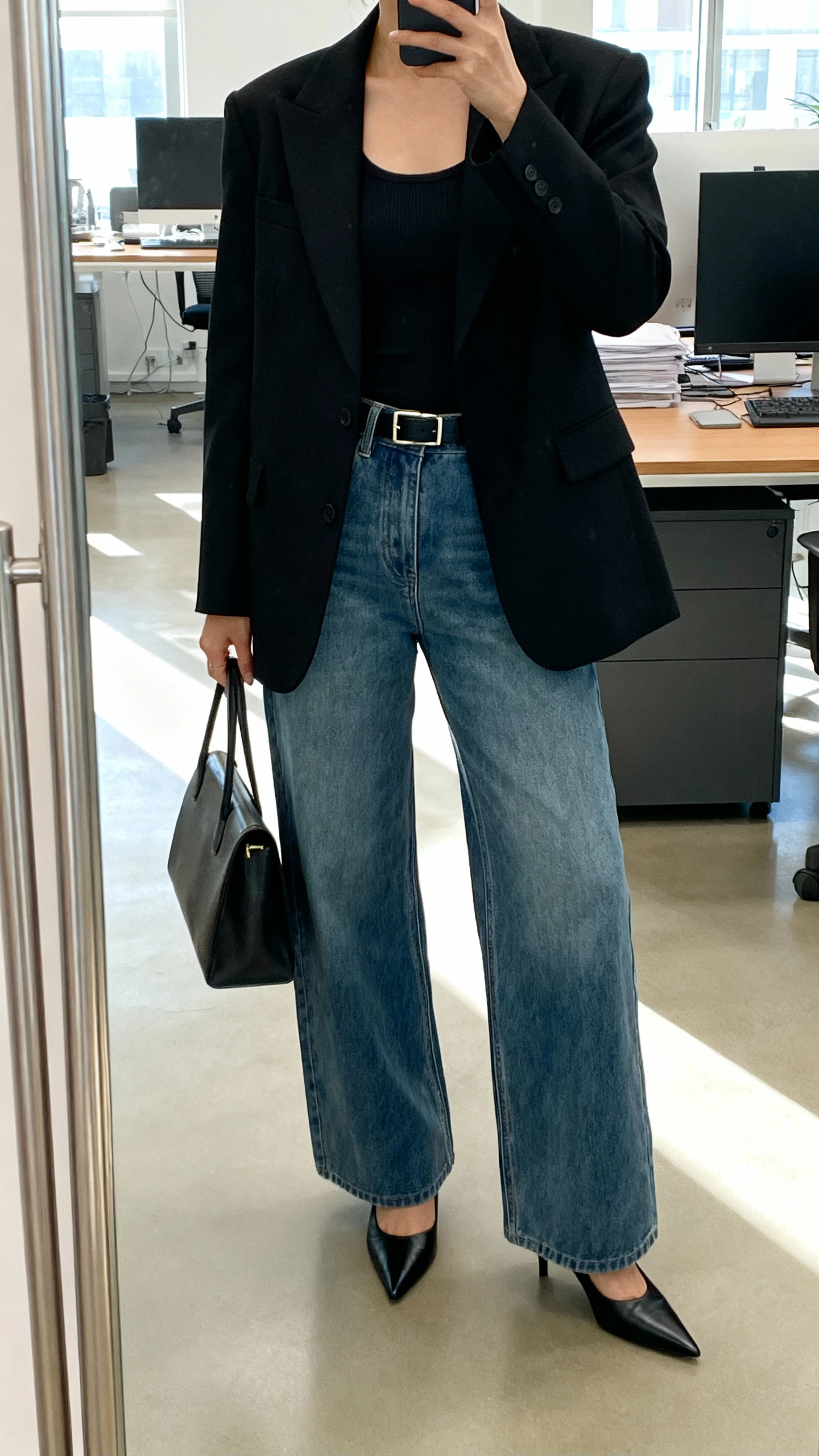 iPhone office mirror selfie of a woman wearing wide leg jeans, a sharp black blazer over a fitted top, black pointed toe heels, structured work bag and a slim belt, face not visible, cropped at shoulders, bright morning office light, natural iPhone photo quality.