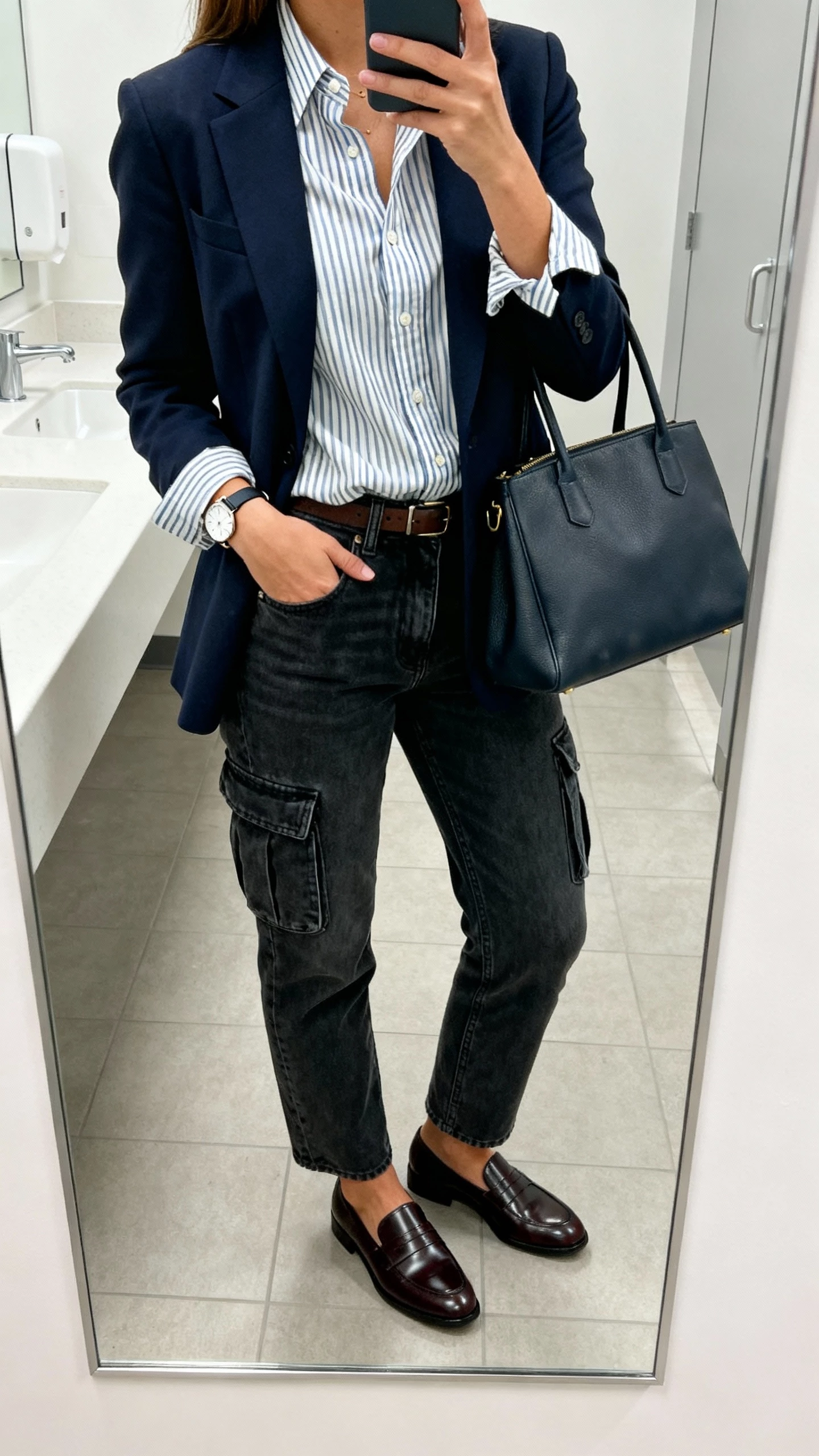 iPhone office restroom mirror selfie of a woman wearing dark-wash cargo jeans, navy blazer, striped button-down shirt half-tucked, polished loafers, structured tote, and a simple watch; face not visible, cropped at shoulders, bright office lighting, realistic iPhone aesthetic.