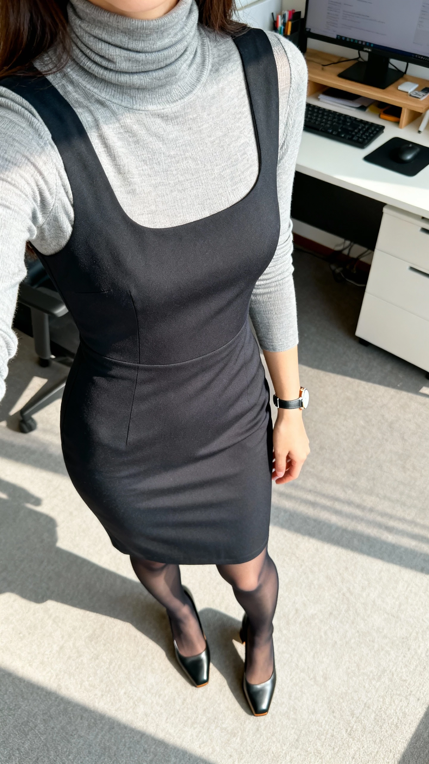 iPhone office selfie cropped at shoulders showing a woman wearing a fitted turtleneck layered under a sheath dress, sheer tights, block-heel pumps, minimal watch, face not visible, desk in background, bright indoor lighting, natural iPhone photo quality.