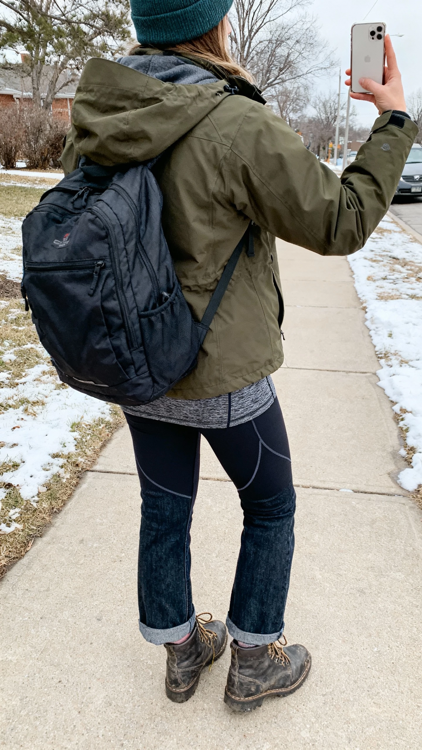 iPhone outdoor back-view selfie of a woman in a forest technical parka with hood, thermal base layer top, heat-tech leggings under thick straight-leg jeans, rugged lace-up boots, water-resistant backpack, merino beanie; snowy sidewalk, face not visible, overcast natural light, casual iPhone photo.