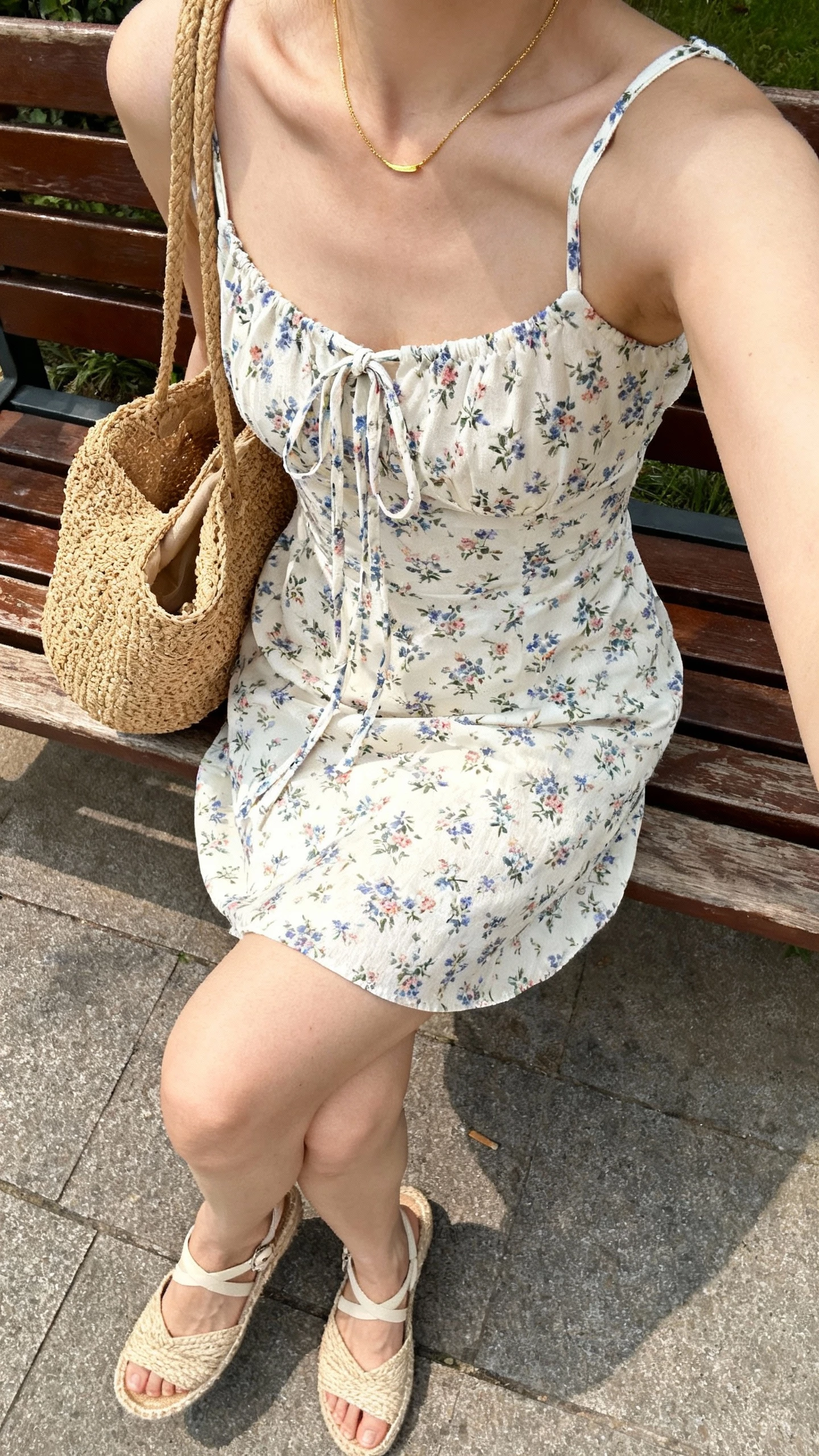iPhone outdoor bench selfie cropped at shoulders showing a woman wearing a daytime floral sundress with tie straps, espadrille wedges, straw tote, and thin gold necklace, face not visible, bright morning light in a park, natural iPhone photo quality