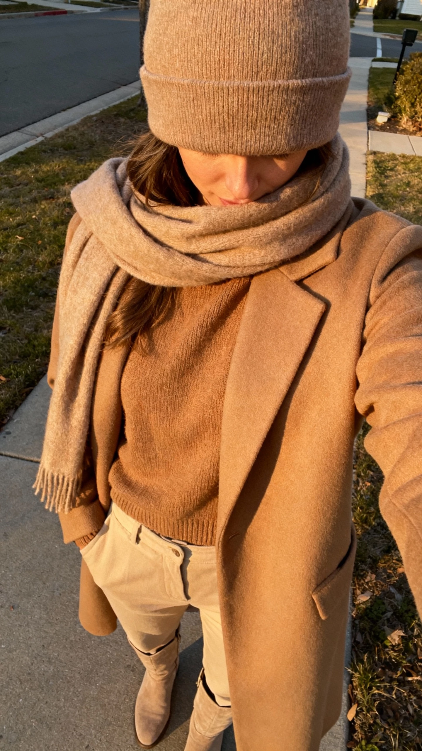 iPhone outdoor selfie cropped at neck showing a woman in a tonal look: beanie, camel coat, tan sweater, beige trousers, sand-colored boots, matching scarf, face not visible, neighborhood sidewalk, golden hour light, natural iPhone photo quality