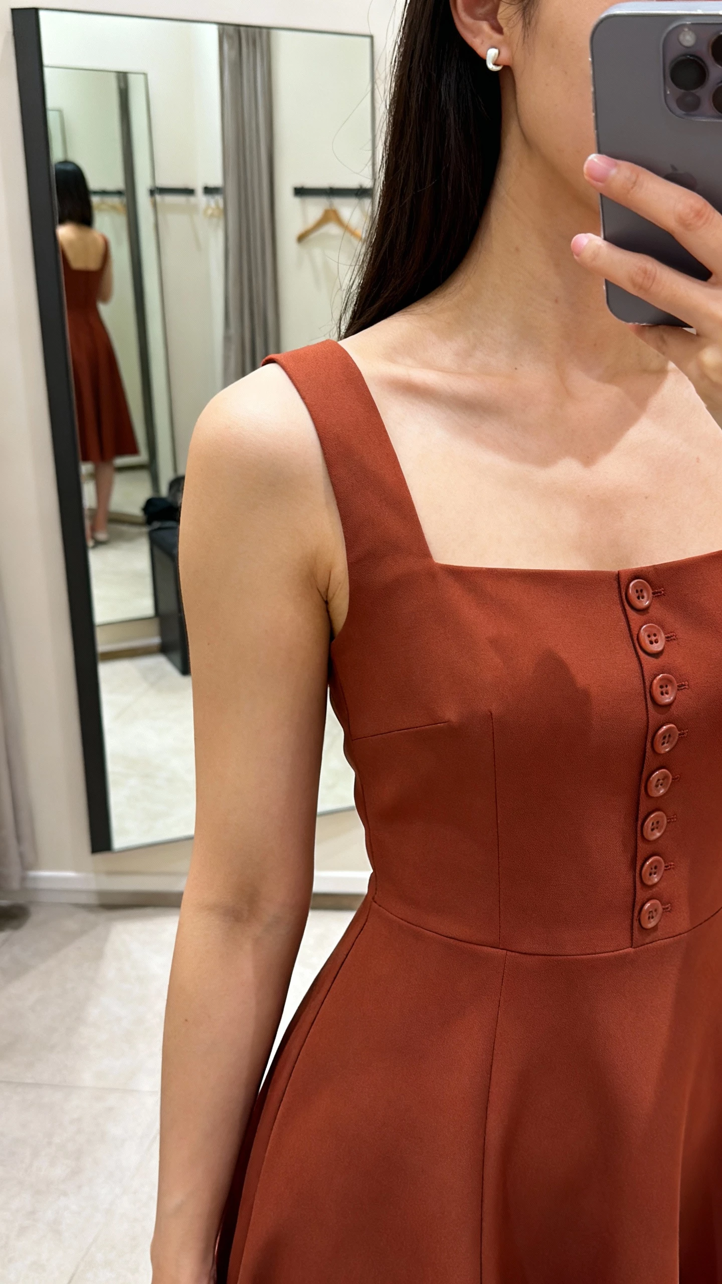 iPhone over-shoulder selfie of a woman in a square-neck crepe fit-and-flare dress with a row of buttons down the back, simple studs, face not visible, dressing room mirror, soft indoor lighting, casual iPhone photo.