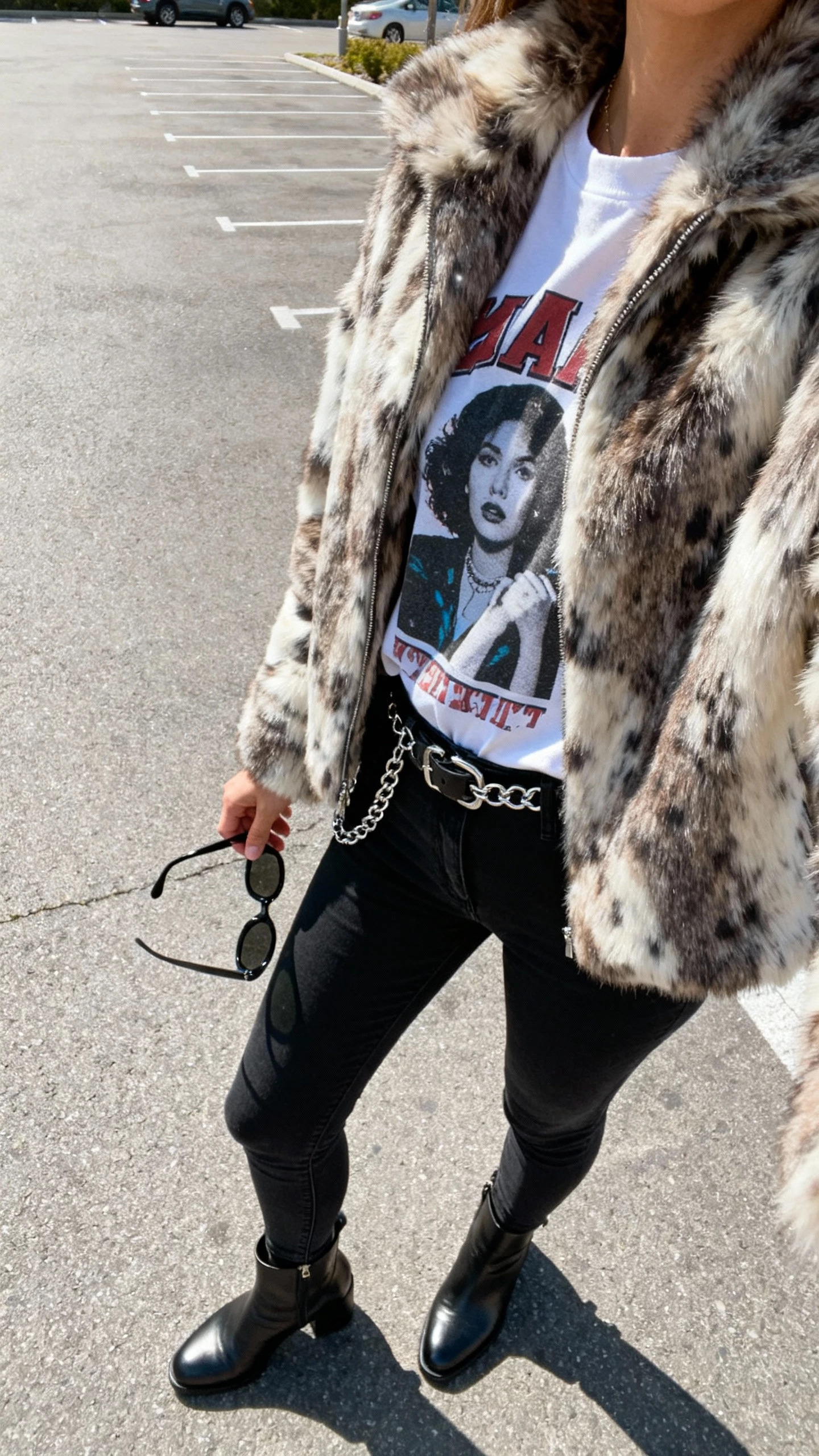 iPhone parking-lot side-angle selfie of a woman in a statement fur jacket over a graphic tee, black skinny jeans, ankle boots, chain belt, sunglasses in hand, face not visible, natural daylight, casual iPhone photo quality.
