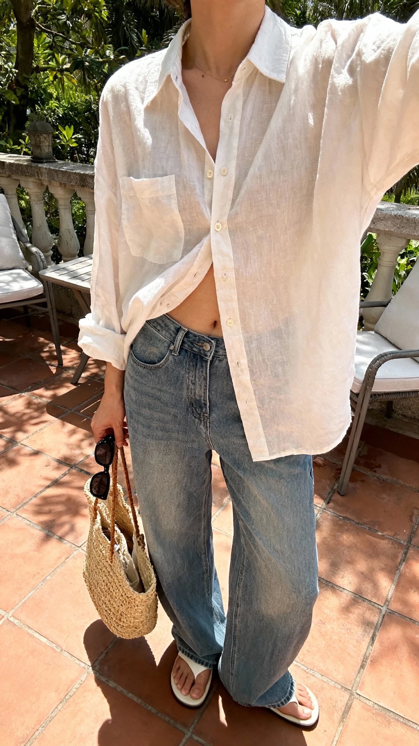 iPhone patio selfie cropped at neck showing a woman wearing wide leg jeans, breezy oversized button-up half-tucked, simple slides, straw tote, sunglasses in hand, face not visible, sunny morning light, natural iPhone photo quality.