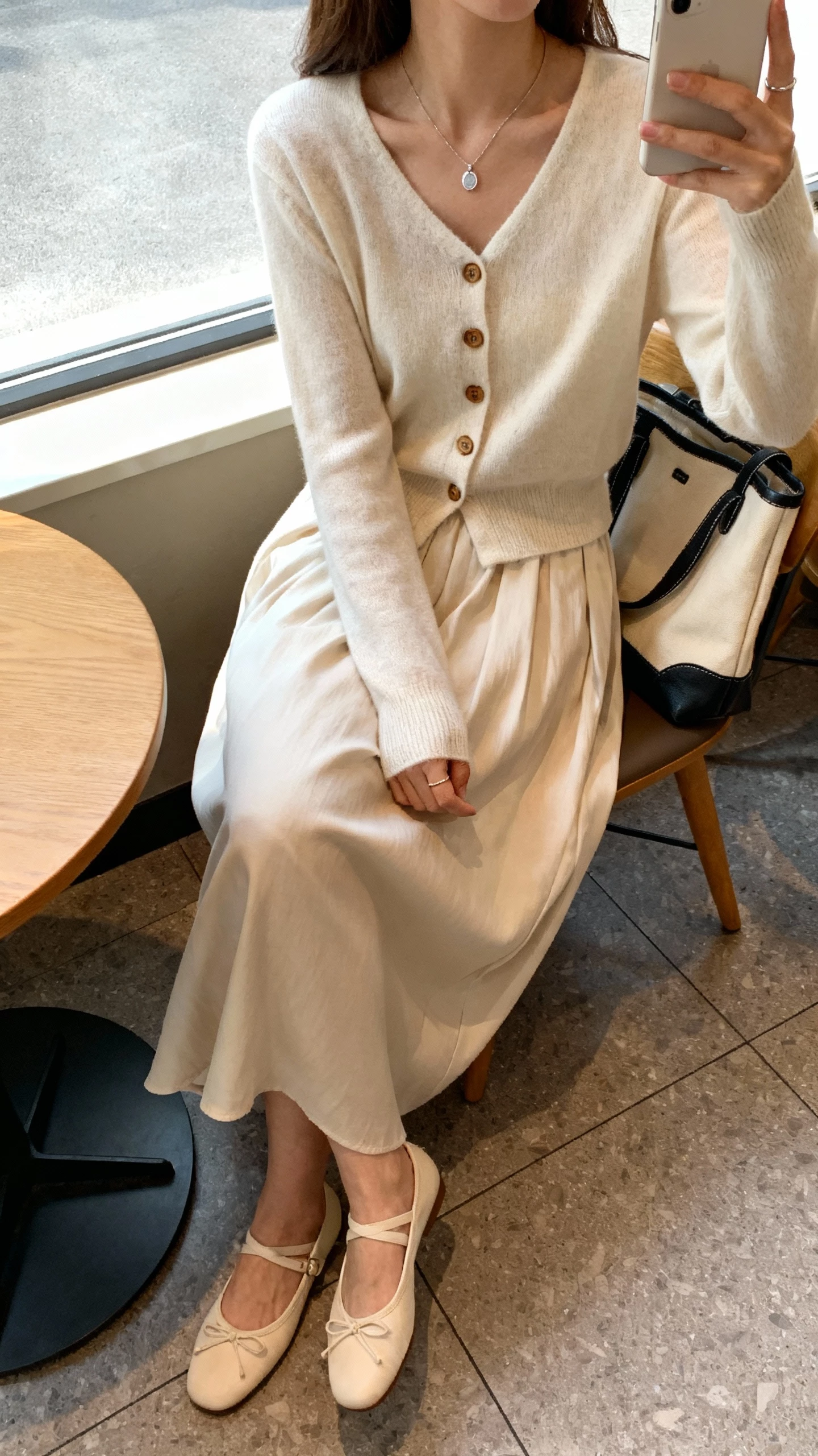 iPhone seated cafe selfie cropped at neck showing a woman in a soft cardigan buttoned at the top with a flowy midi skirt, ballet flats, delicate pendant necklace, tote on chair, face not visible, window light, natural iPhone photo quality.