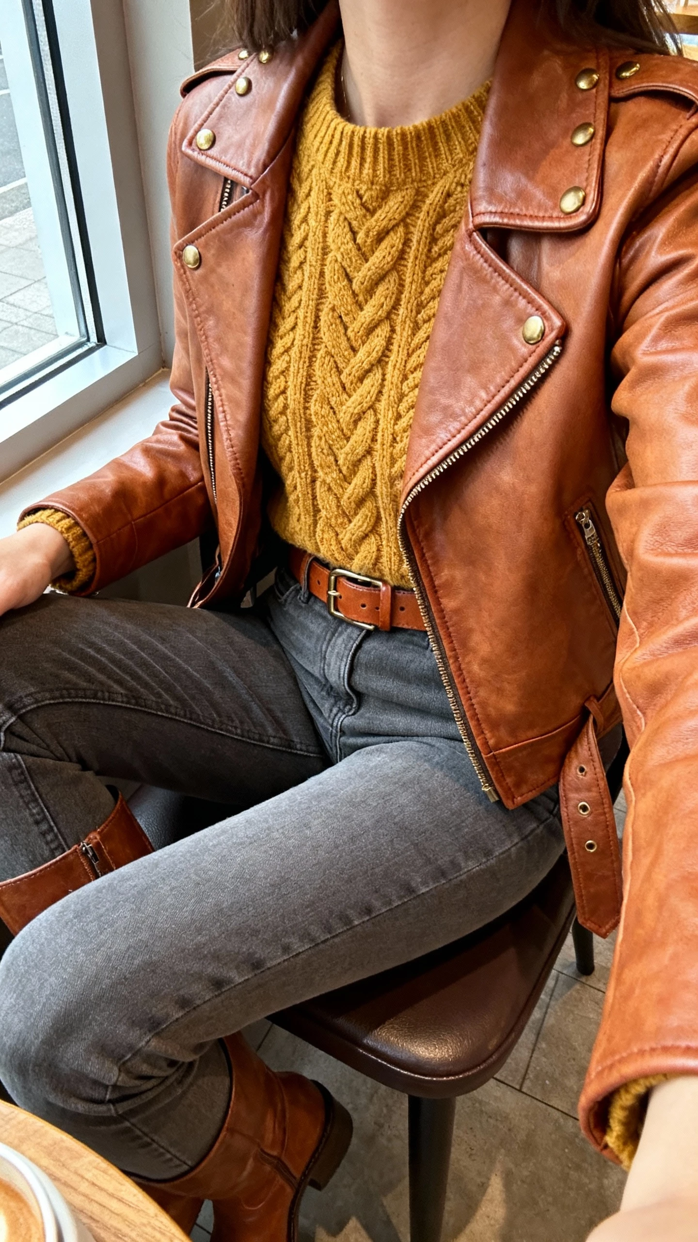 iPhone seated cafe selfie cropped at neck showing a woman wearing a mustard cable-knit sweater, slate gray straight-leg jeans, warm brown leather moto jacket, cognac belt and boots, brass studs, face not visible, window light, natural iPhone photo
