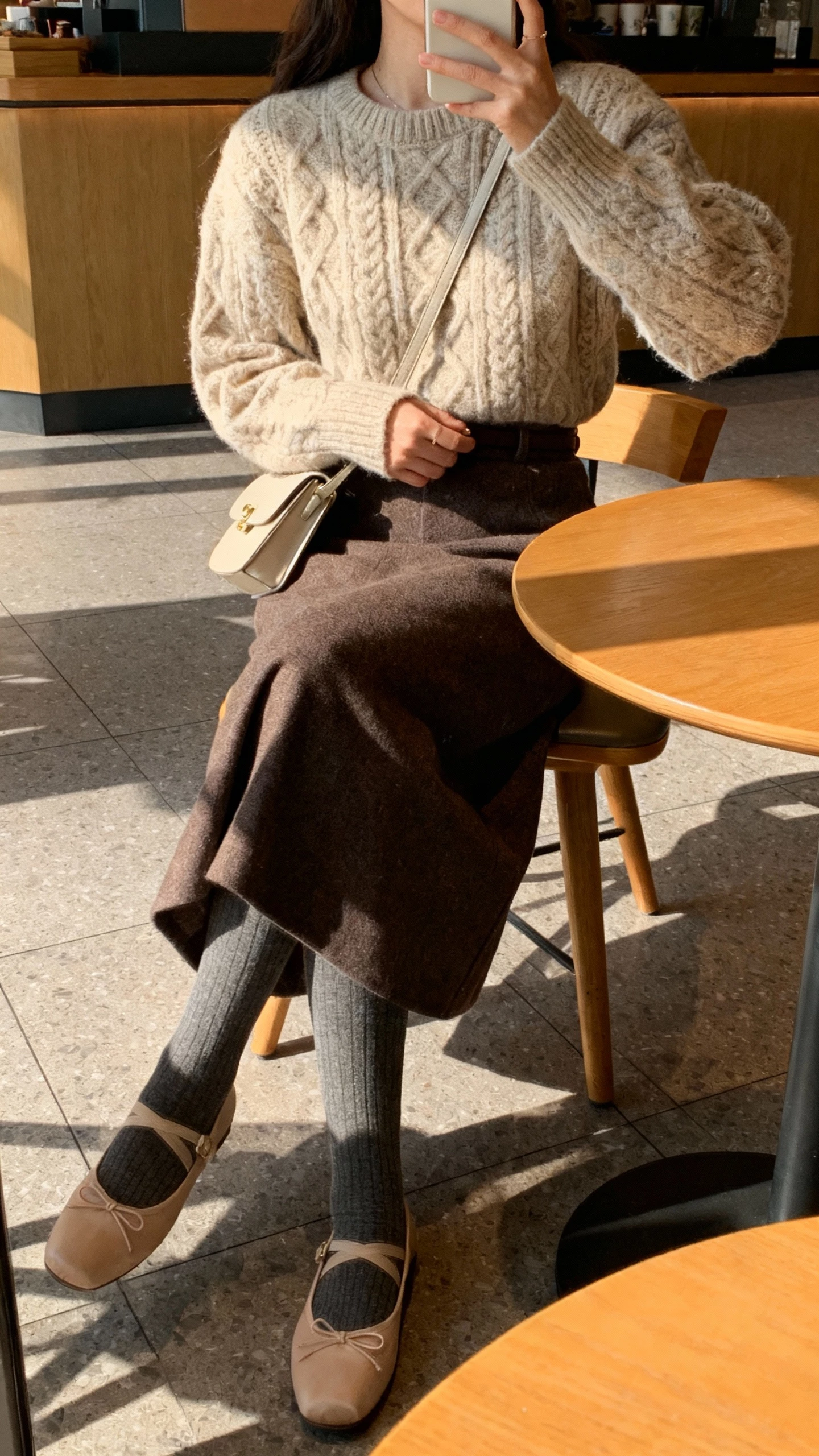 iPhone seated cafe selfie cropped at neck showing a woman in a cozy knit sweater tucked into a midi skirt with tights, ballet flats, and a small shoulder bag, face not visible, warm afternoon light, natural iPhone quality.