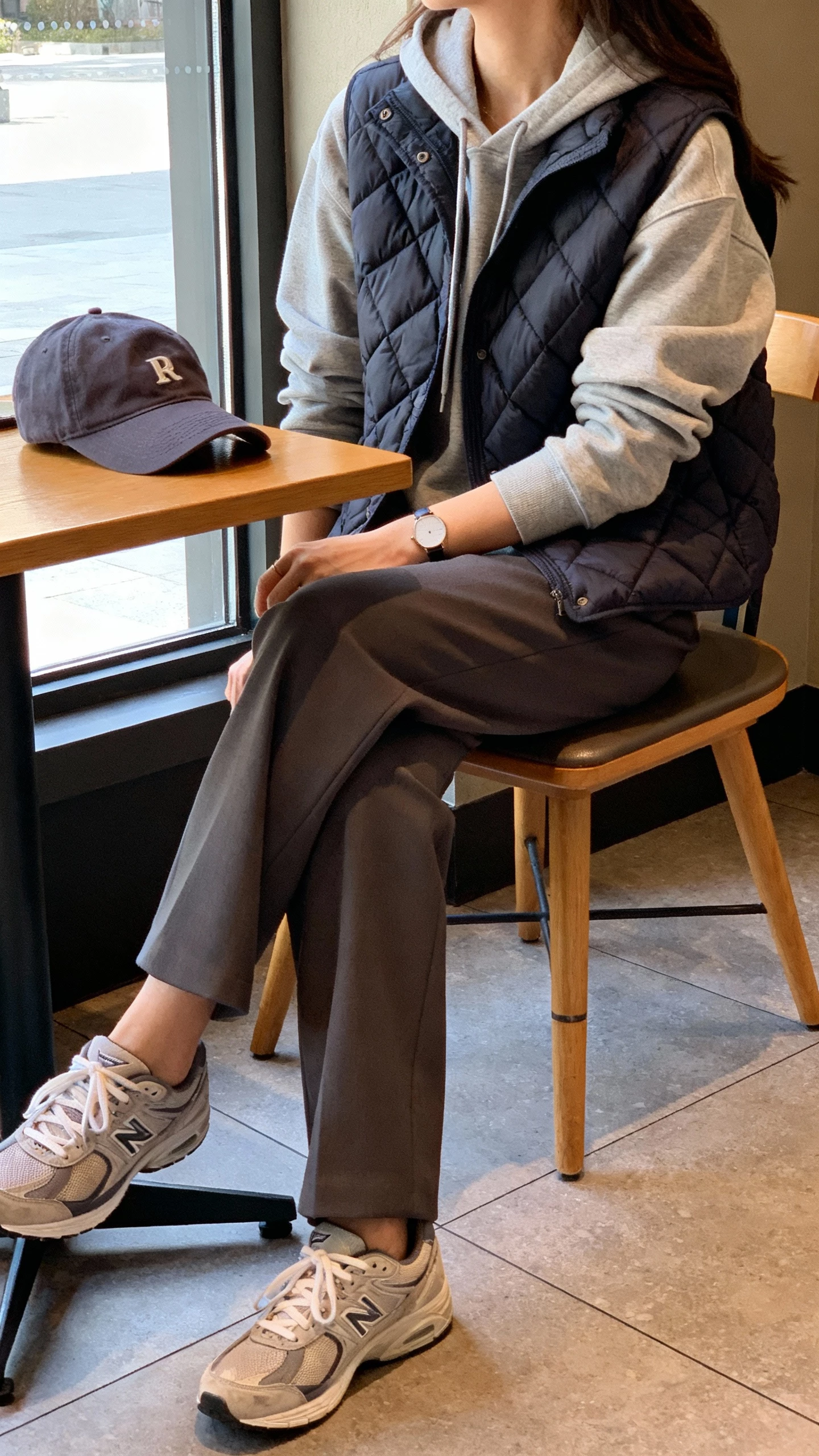 iPhone seated cafe selfie cropped at shoulders of a woman wearing a quilted vest over a hoodie, tailored trousers, clean sneakers, baseball cap tucked beside her, and a minimalist watch, face not visible, window light, casual iPhone aesthetic.