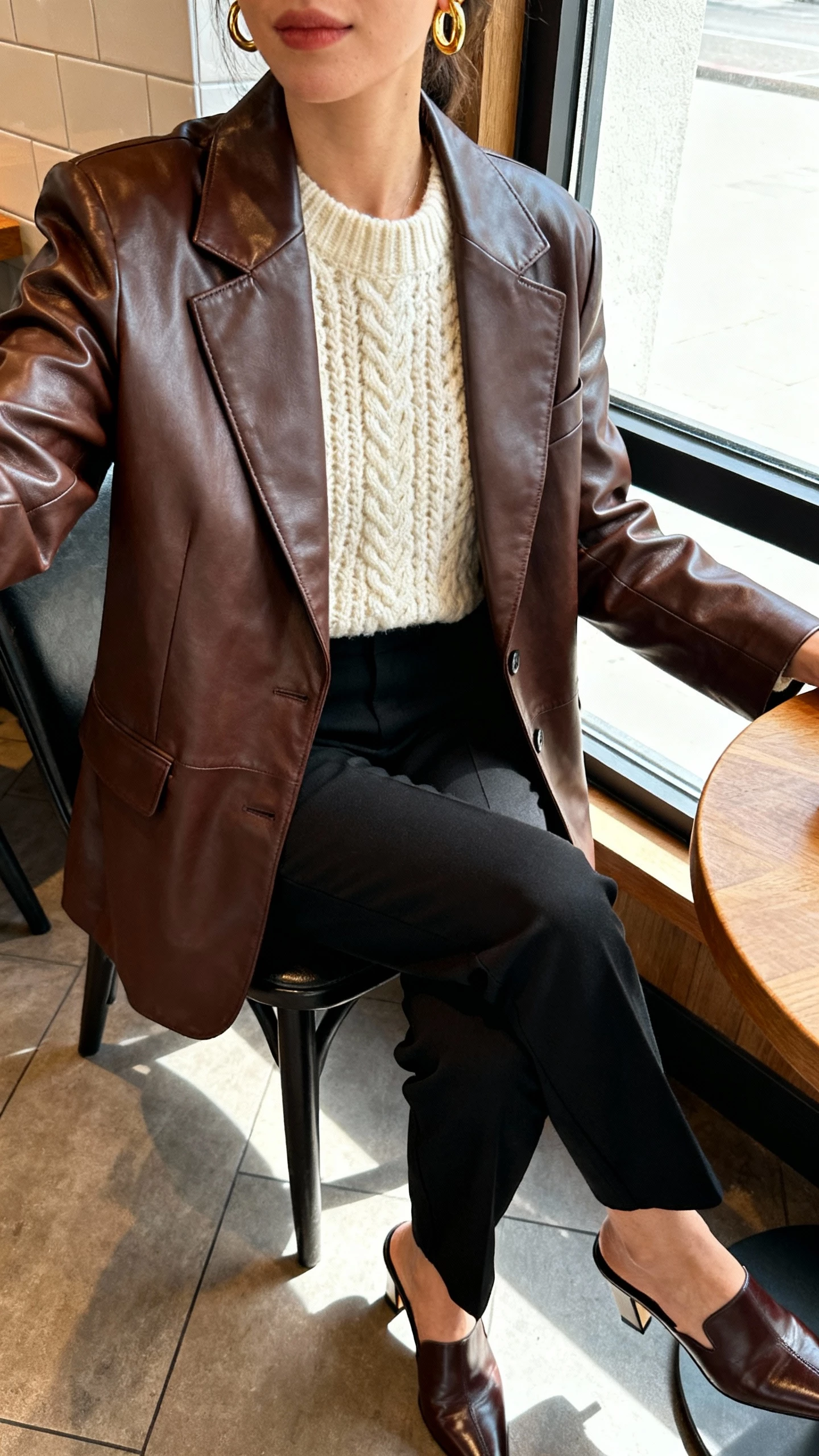 iPhone seated cafe selfie of a woman in luxe leather + knit contrast: chocolate leather blazer over a cream chunky knit, black tailored pants, heeled leather mules, and gold hoop earrings, face not visible, cropped at shoulders with window light, natural iPhone photo.